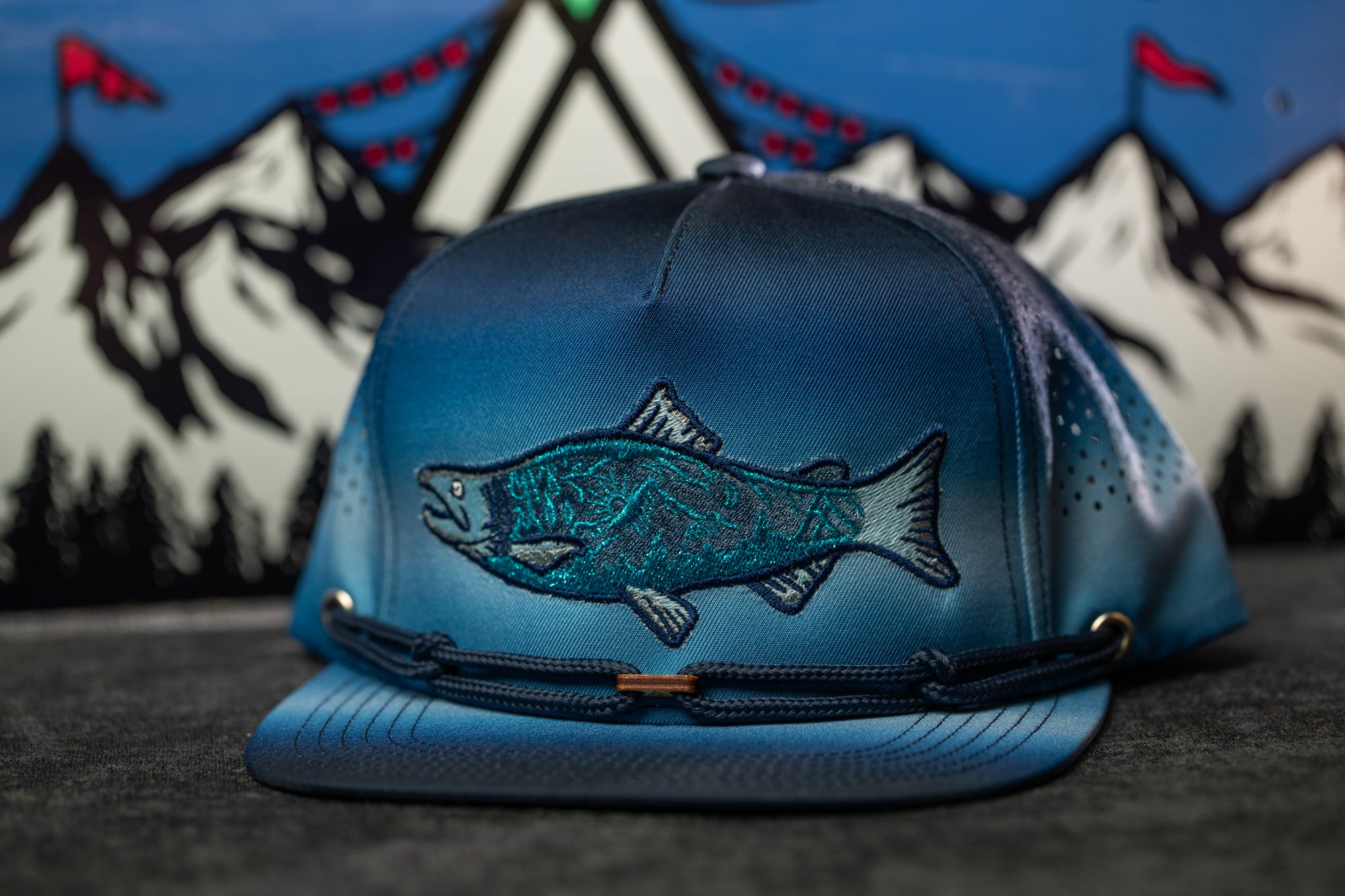 A blue baseball cap with a fish embroidered on it, placed on a surface with a mountain landscape in the background.