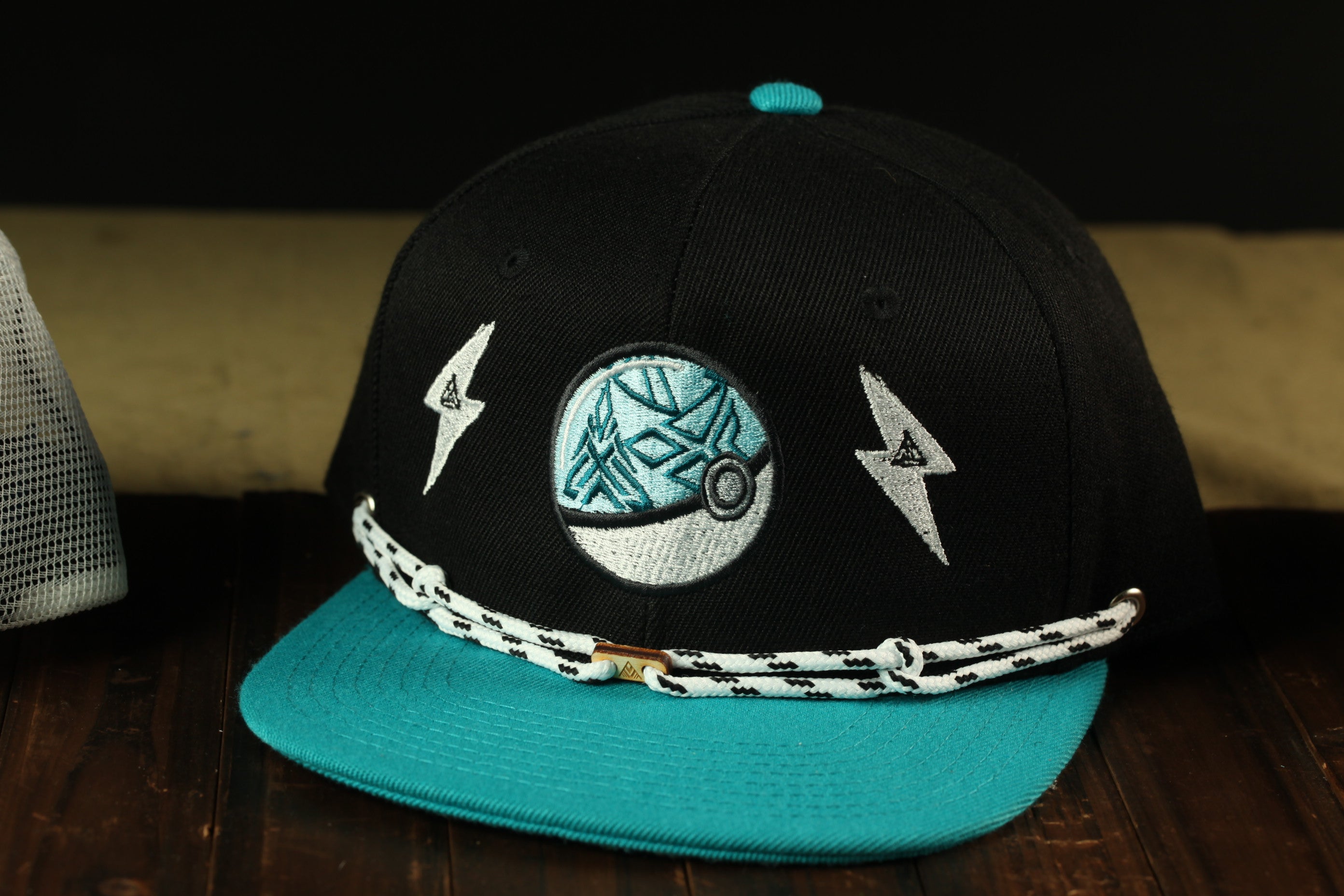 A black baseball cap with a teal brim and a white lightning bolt design on the front, placed on a wooden surface.