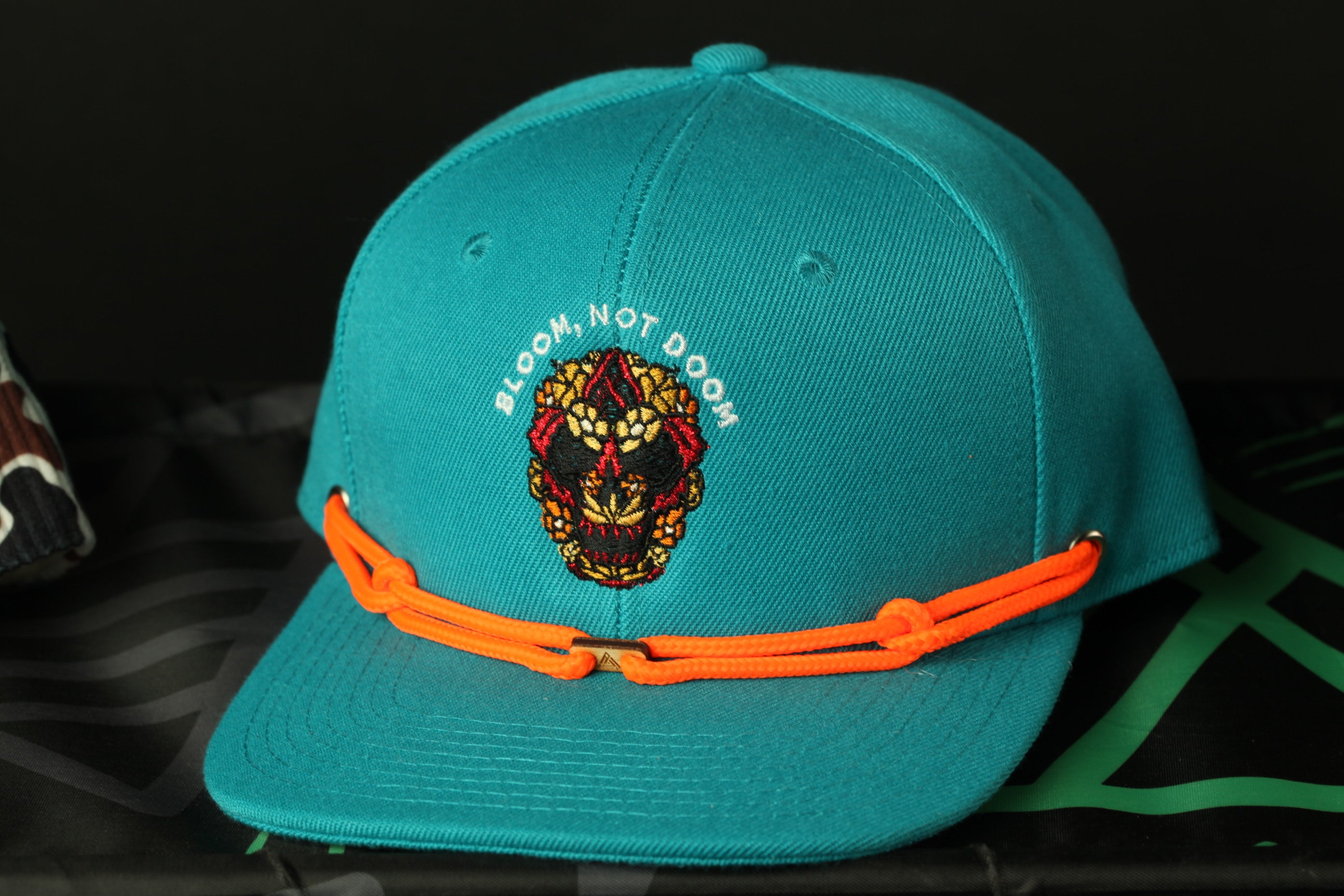 A turquoise baseball cap with an orange string and a skull design on the front.