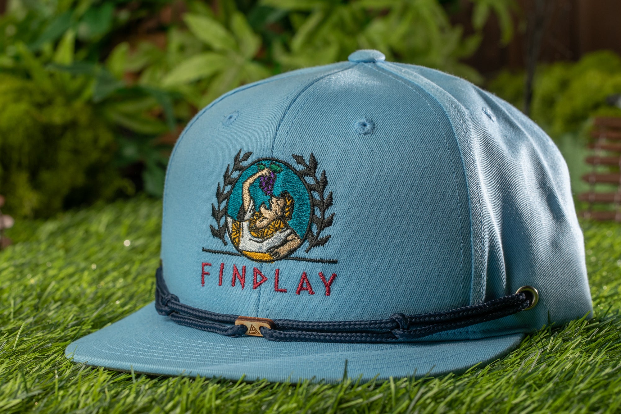 A light blue baseball cap with a graphic design and the word "FINDLAY" printed on it, sitting on a grassy surface.