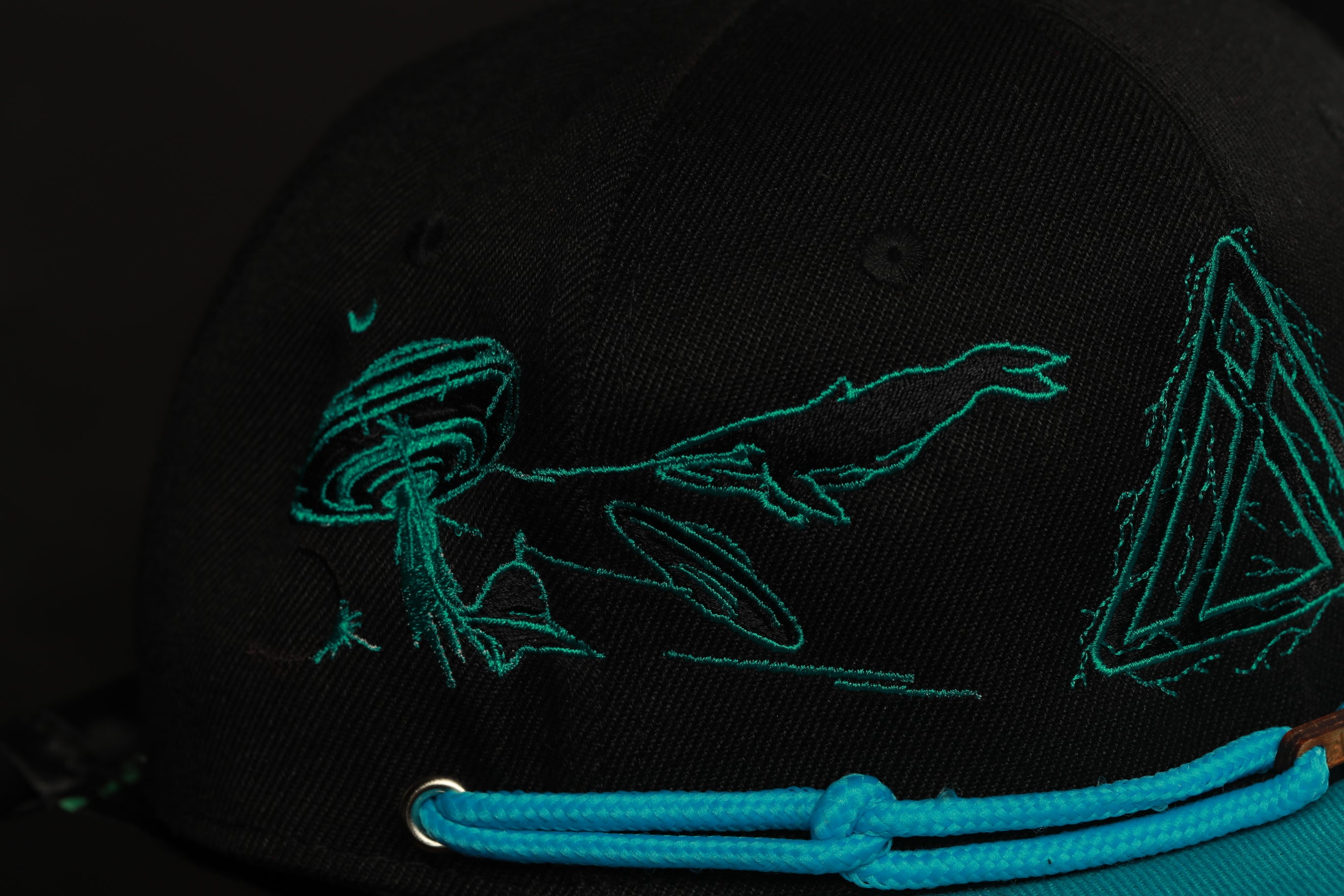 a close-up view of a black baseball cap with a unique embroidered design featuring a green alien-like creature and a geometric pattern.
