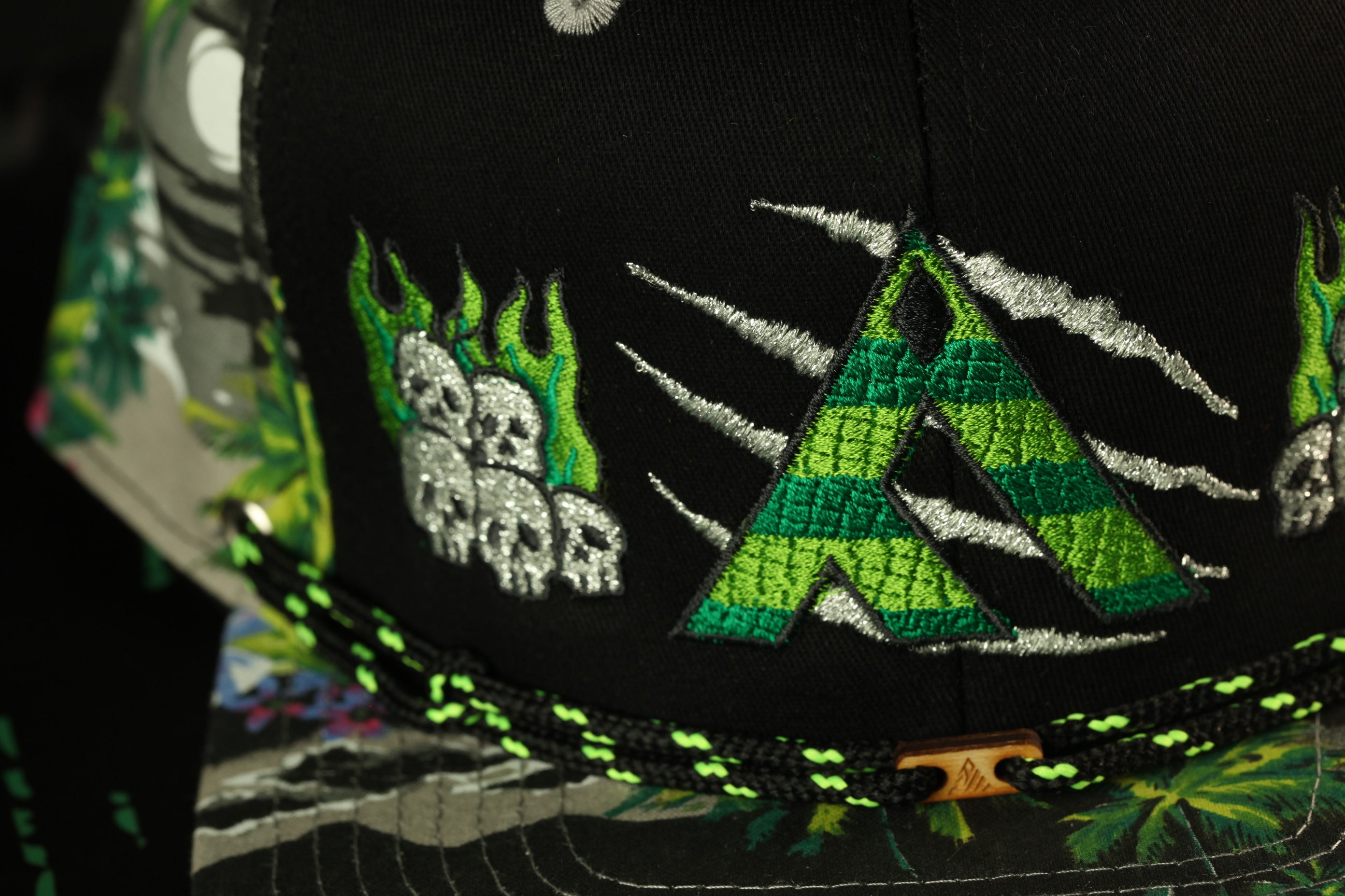 a close-up view of a black baseball cap with various colorful and intricate embroidered designs, including a large green and black "A" and other patterns.