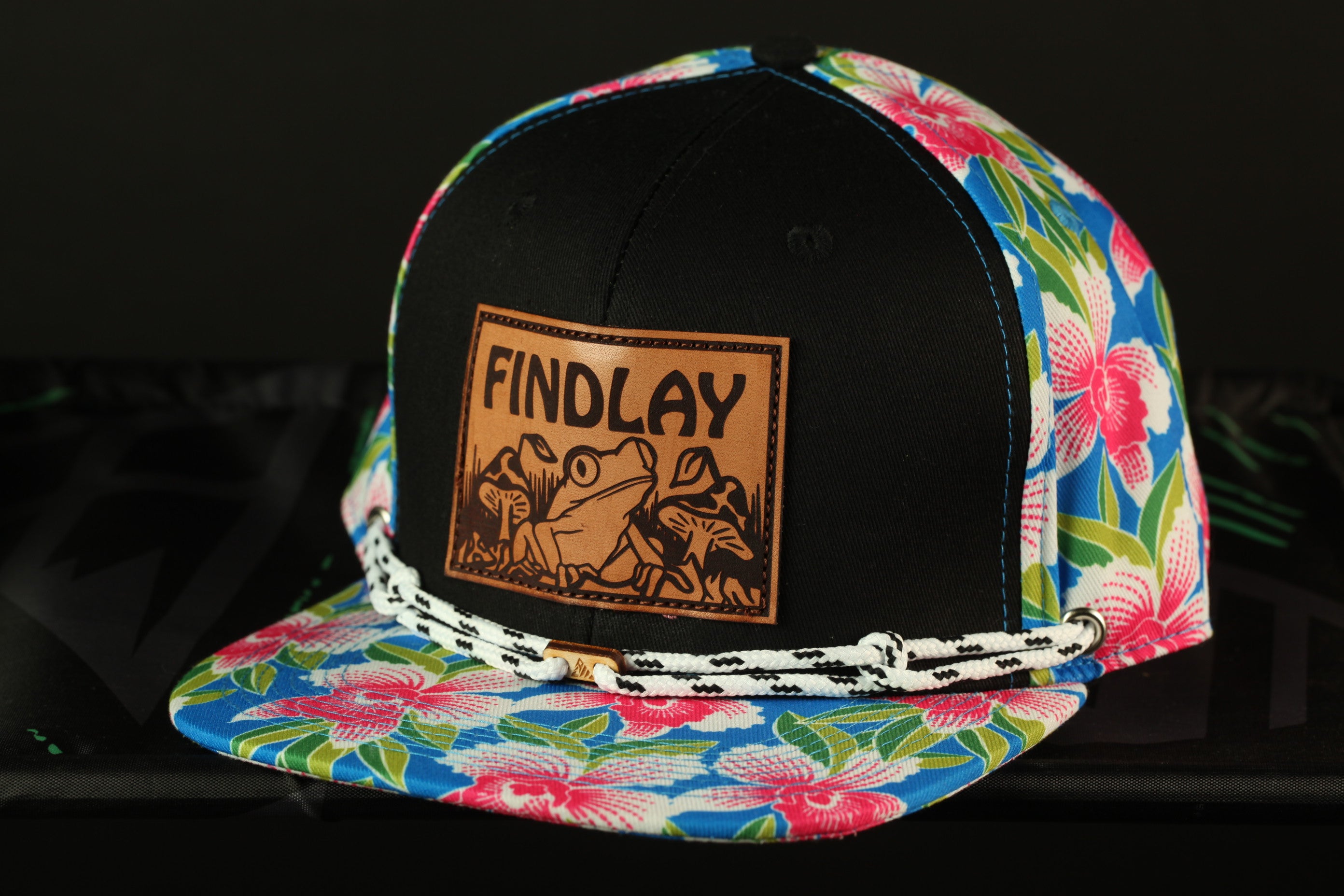 A colorful baseball cap with a black brim and a floral pattern on the front. The cap has a leather patch on the front that says "FINDLAY".