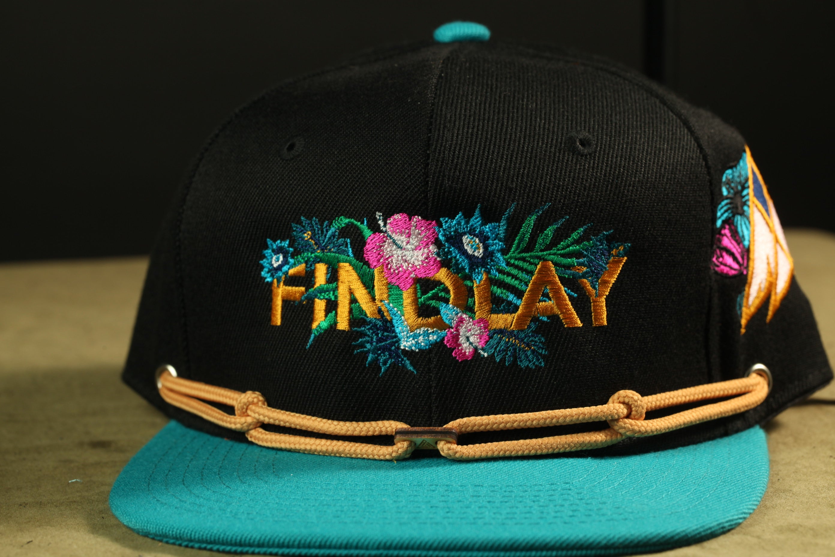 A black baseball cap with a teal brim and a floral embroidered logo on the front.
