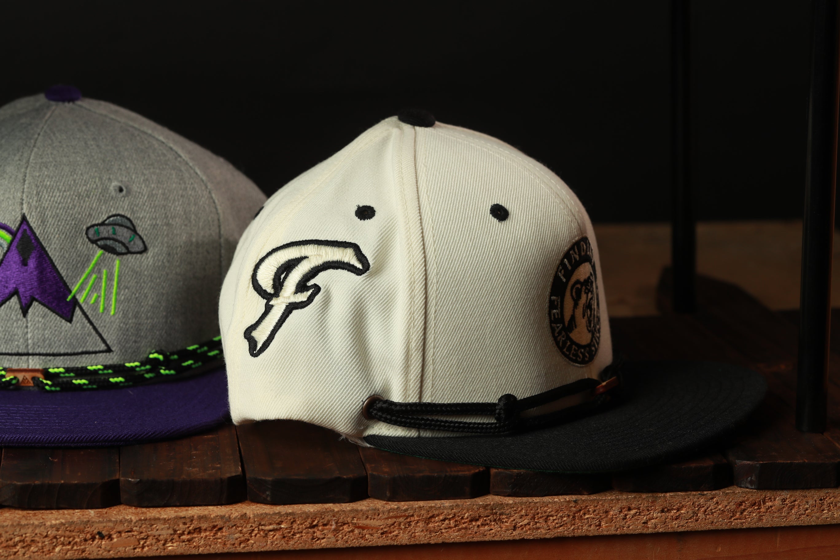 Two baseball caps are displayed on a wooden surface, one white and one gray, with a black strap.