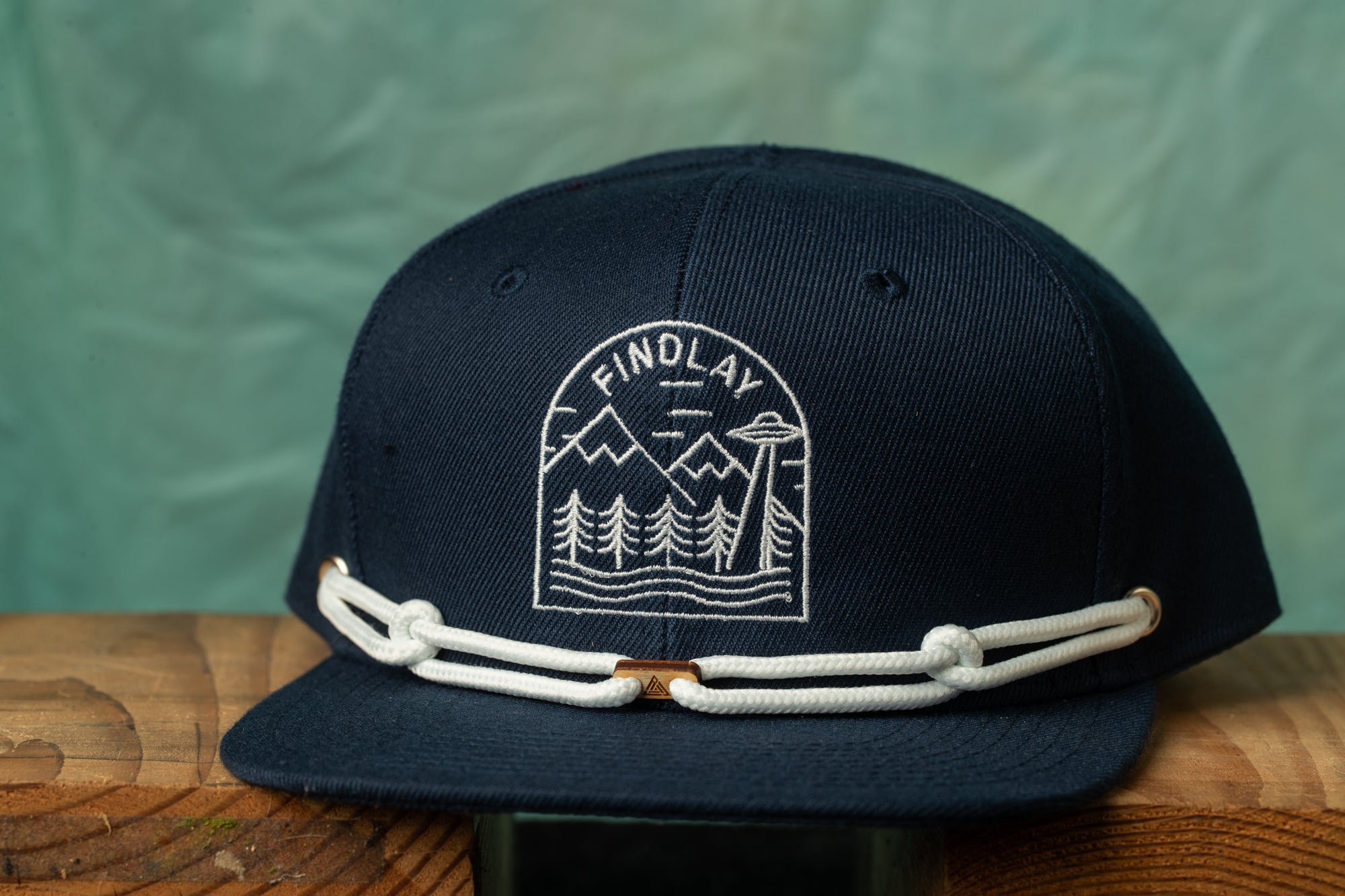 A navy blue baseball cap with a white graphic design on the front, featuring a mountain landscape and an alien spacecraft.