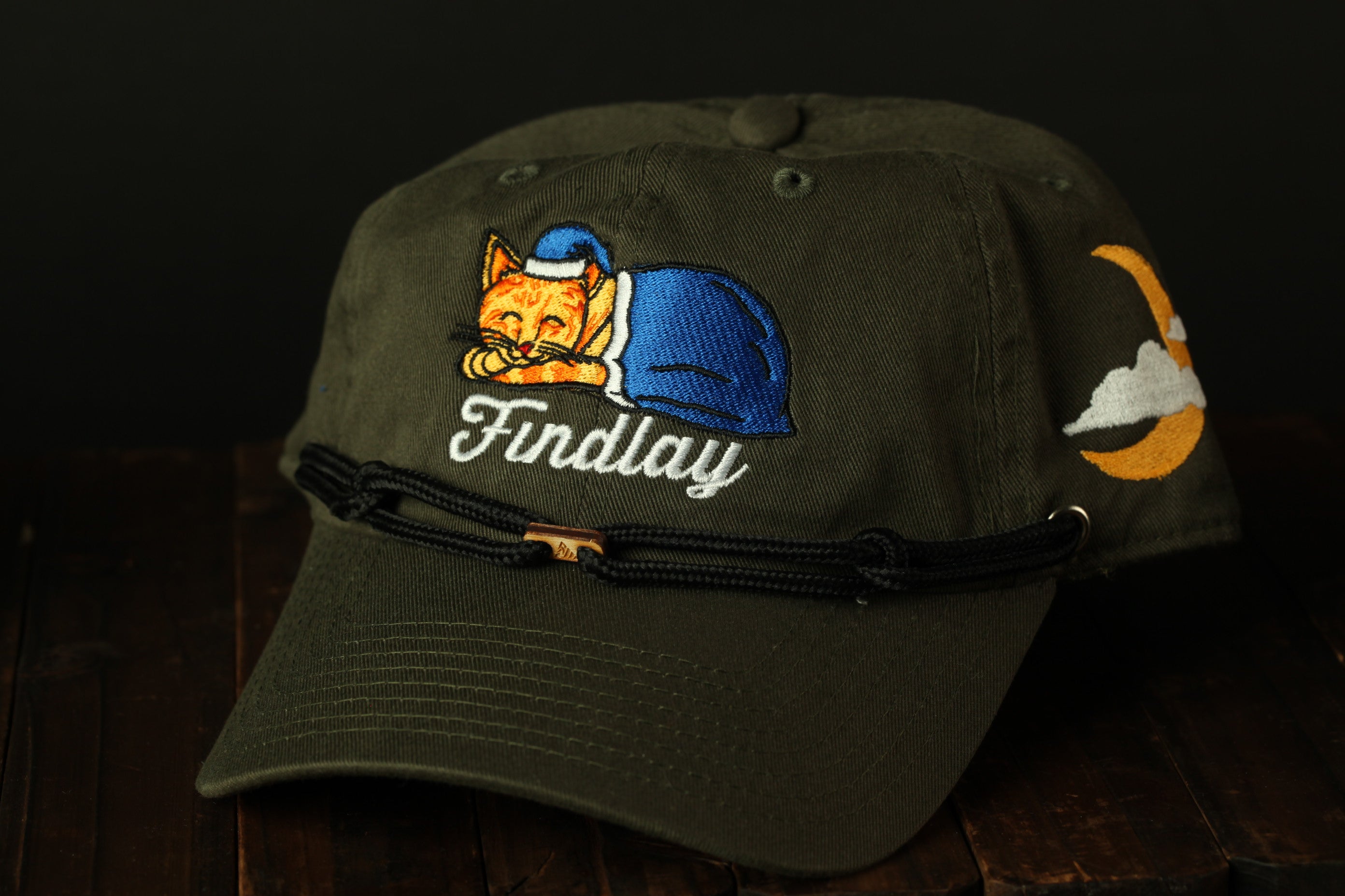 A dark green baseball cap with a cat embroidered on it, featuring a blue blanket and a crescent moon.