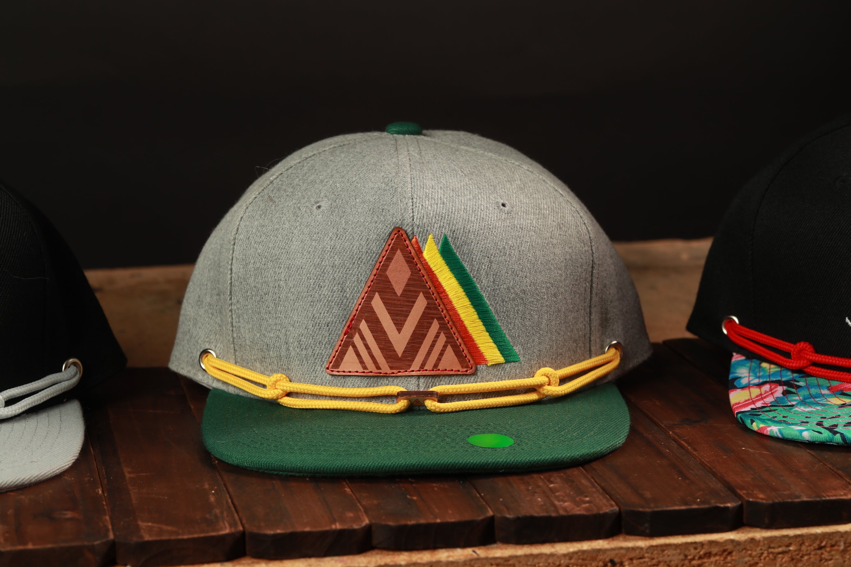 A gray baseball cap with a triangular patch and yellow straps is displayed on a wooden surface, with other hats visible in the background.