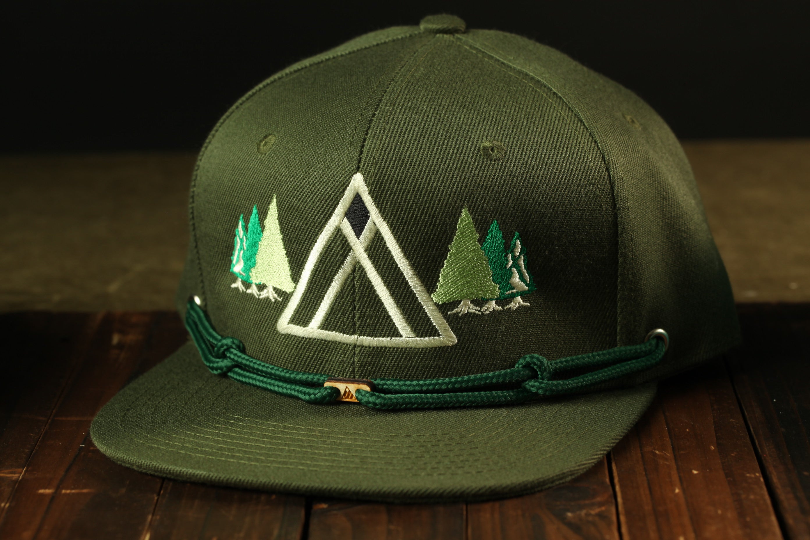 A green baseball cap with a white triangle design and embroidered pine trees on the front, sitting on a wooden surface.