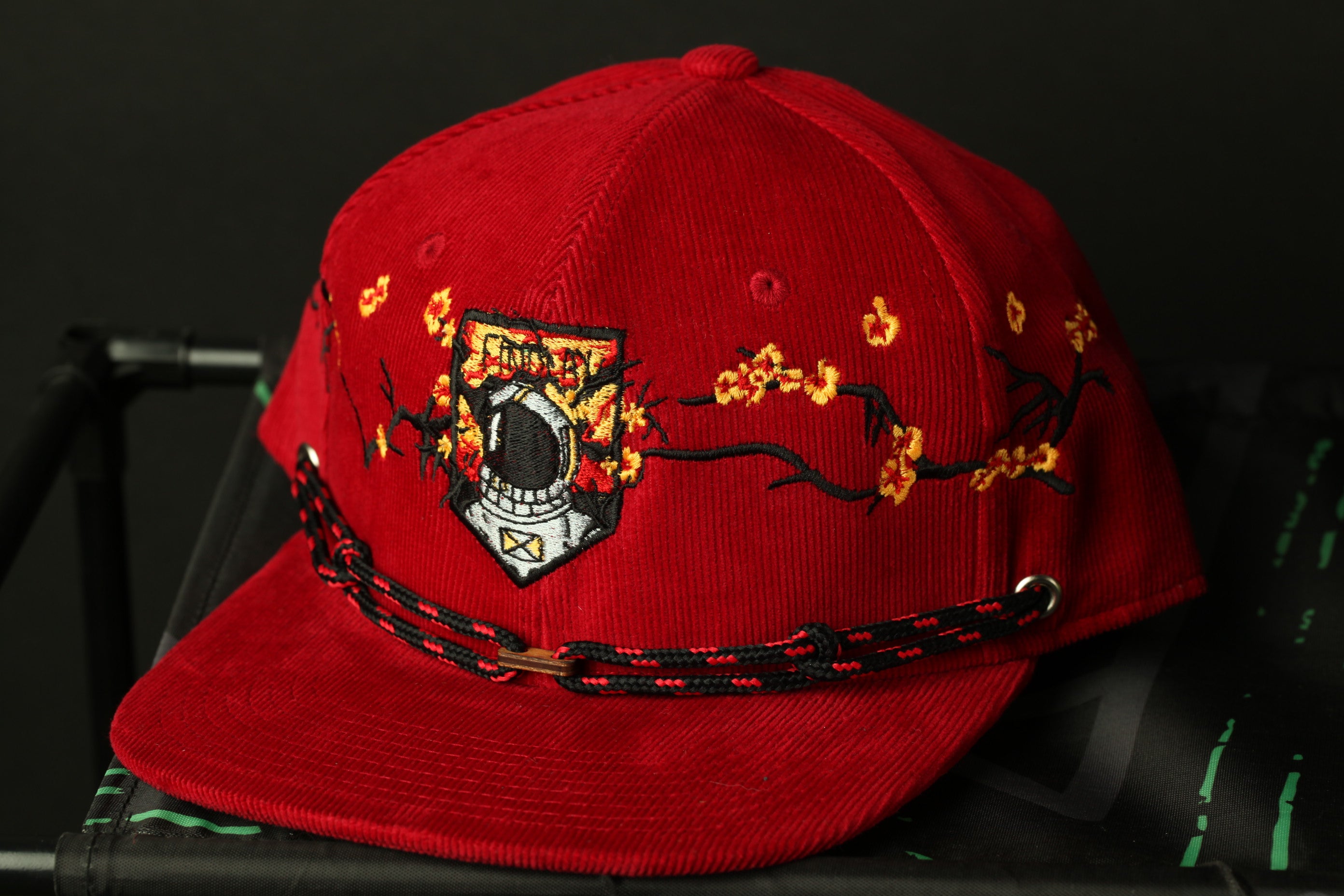 A red baseball cap with a black and red braided band and a white and black embroidered logo on the front.