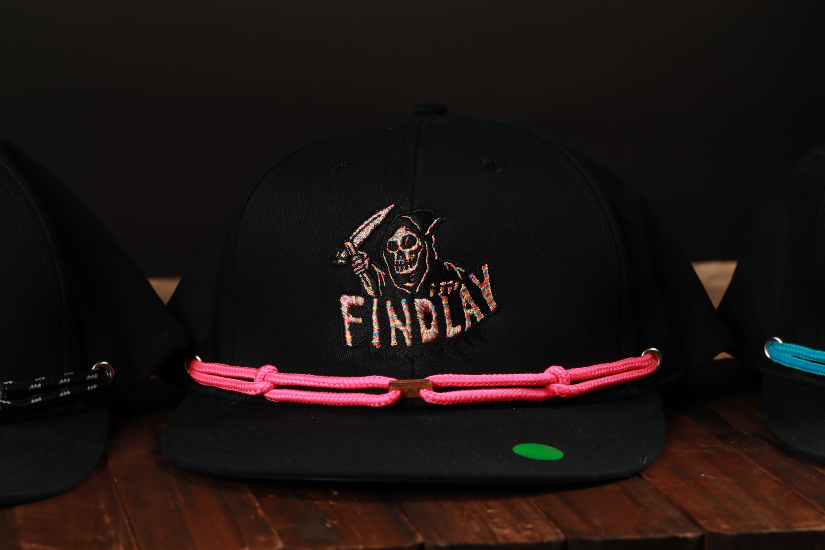 A black baseball cap with a pink braided strap and a skull design on the front, displayed on a wooden surface.