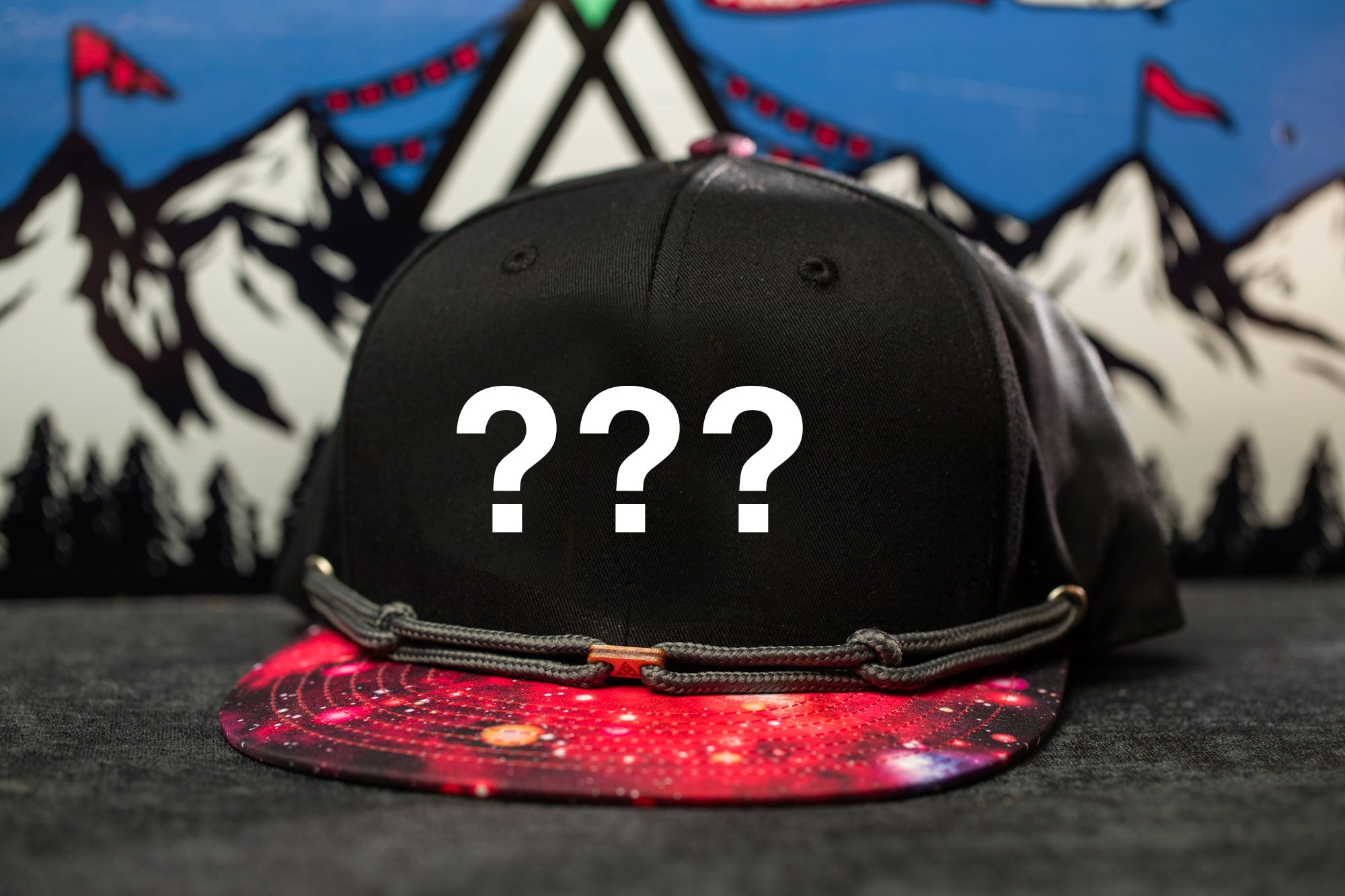 A black baseball cap with a colorful pattern and three question marks printed on it, placed on a surface with a mountain landscape in the background.