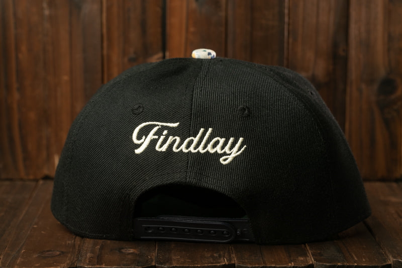 A black baseball cap with the word "Findlay" printed on it, sitting on a wooden surface.