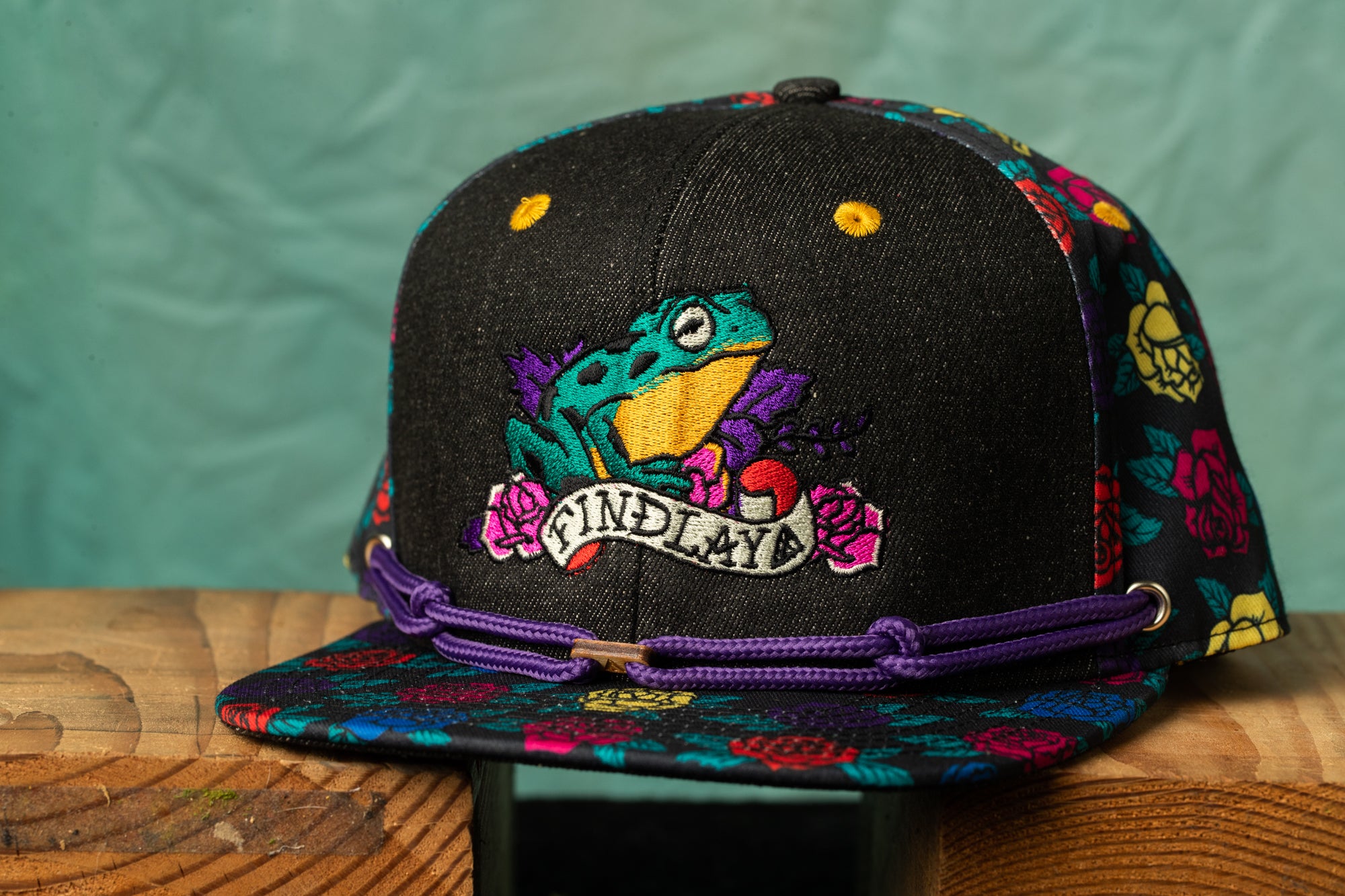 A colorful baseball cap with a frog design and the word "FINDLAYA" printed on it, sitting on a wooden surface.