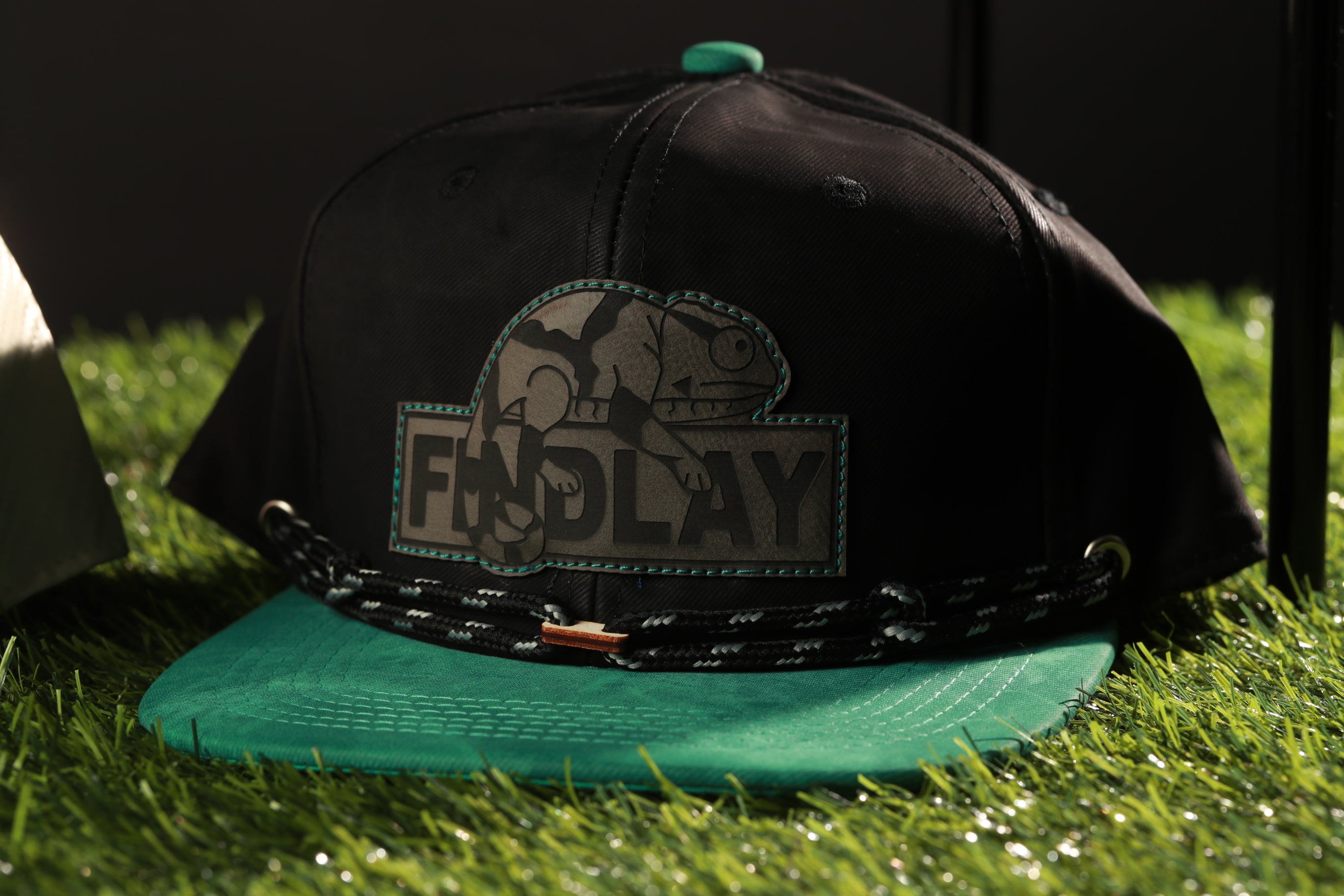 A black baseball cap with a green brim and a black and teal logo on the front, placed on a grassy surface.