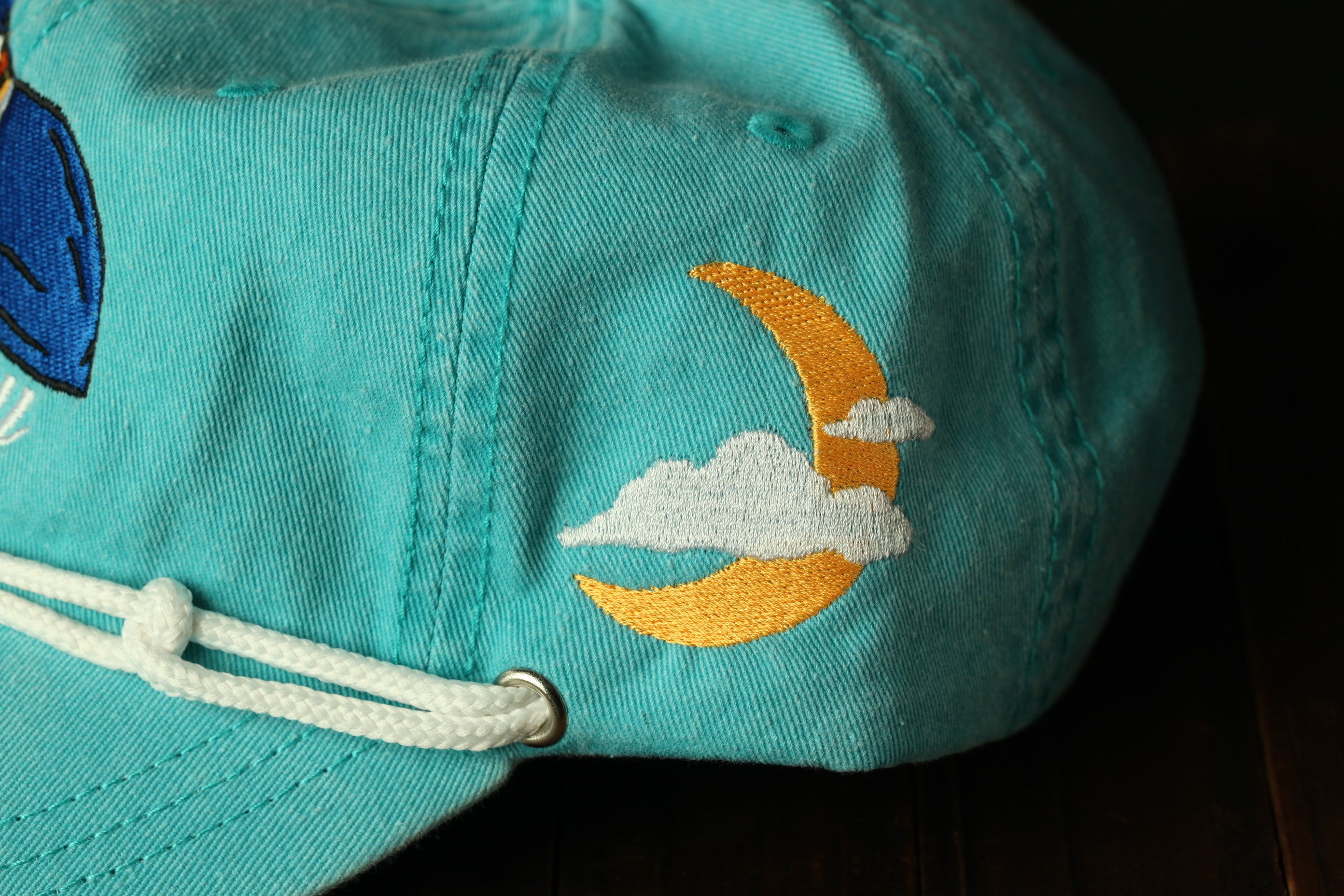 A close-up view of a turquoise baseball cap with a crescent moon and clouds embroidered on it.