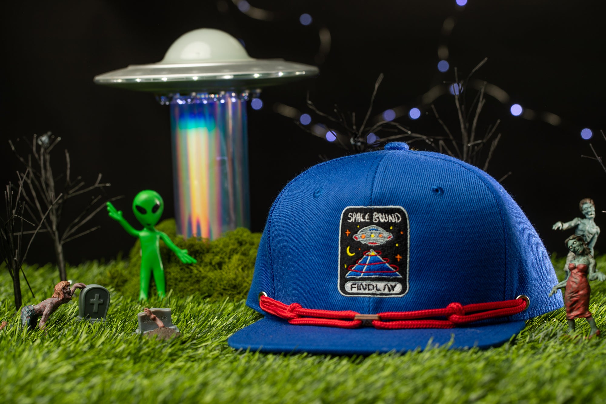 a blue baseball cap with a patch on it, surrounded by various objects such as a green alien figurine, a skull, and a spaceship.