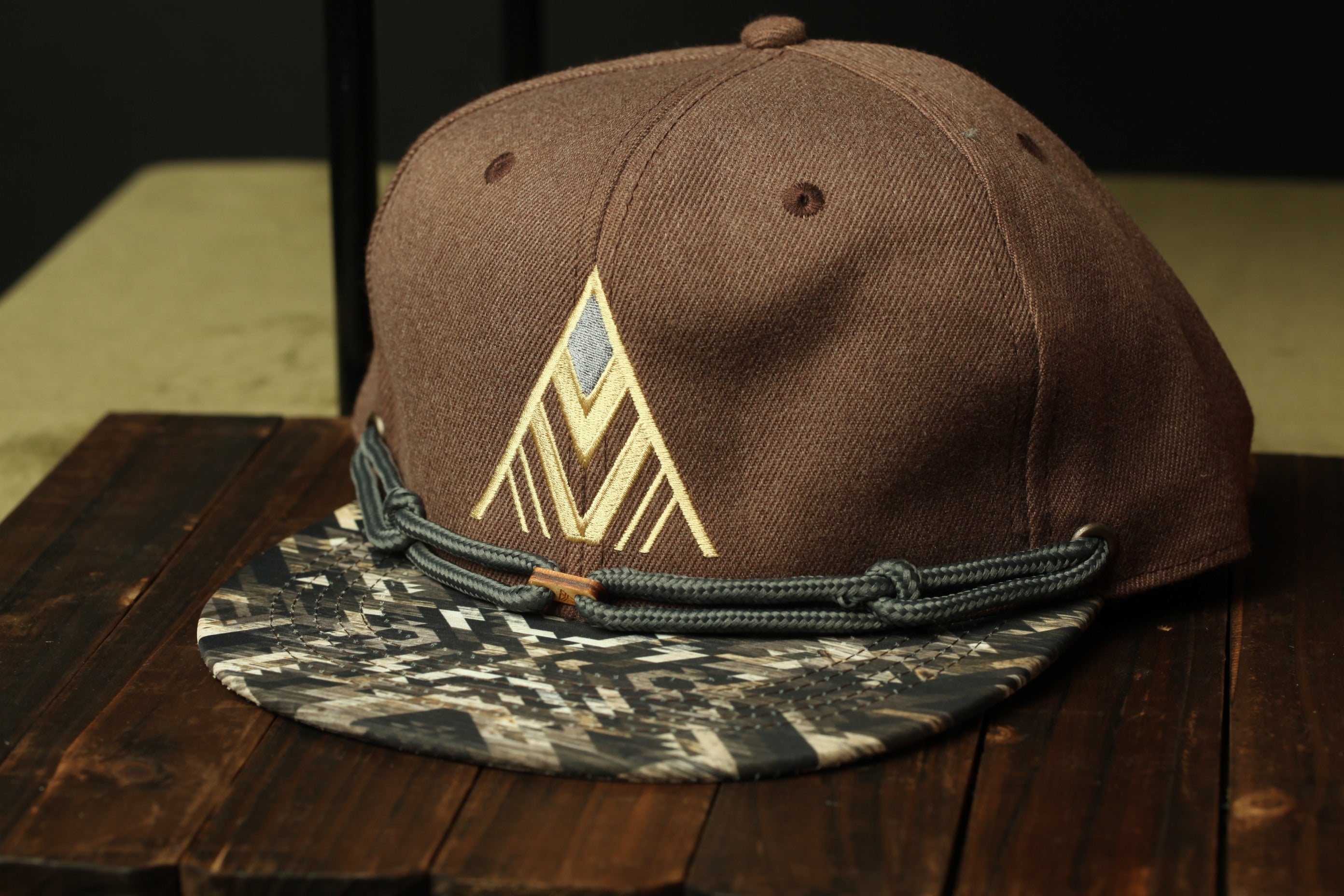 A brown baseball cap with a geometric pattern and a black strap is displayed on a wooden surface.