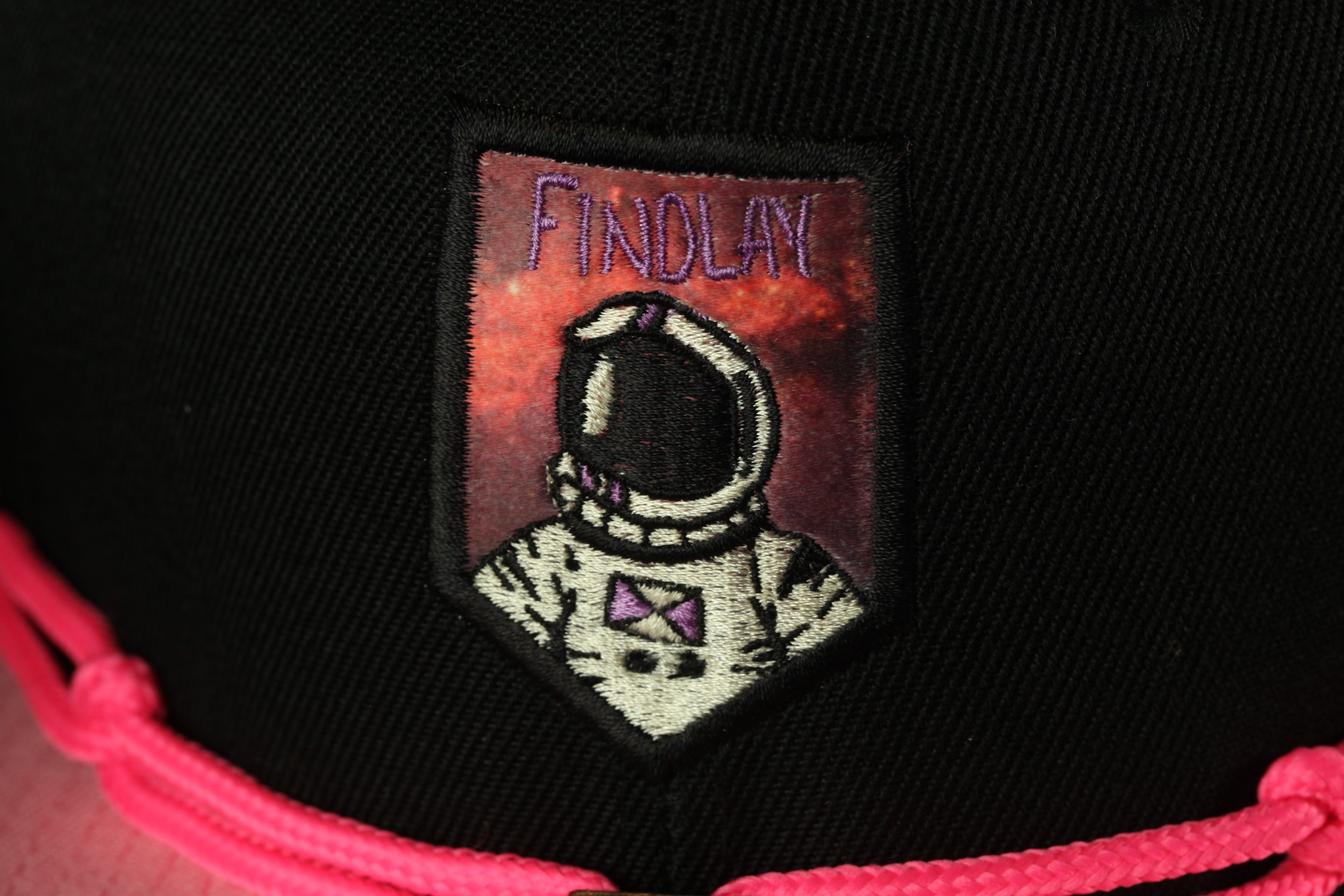 a close-up of a black baseball cap with a patch on it, depicting an astronaut in a white suit with a purple helmet.