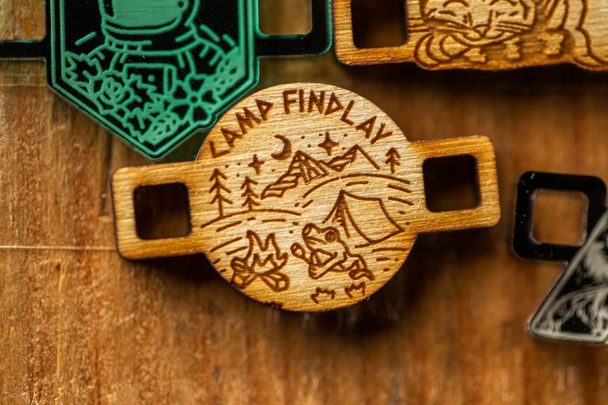 a wooden charm or bracelet with a carved design, including a camping scene and the text "LAMP FIND LAY".