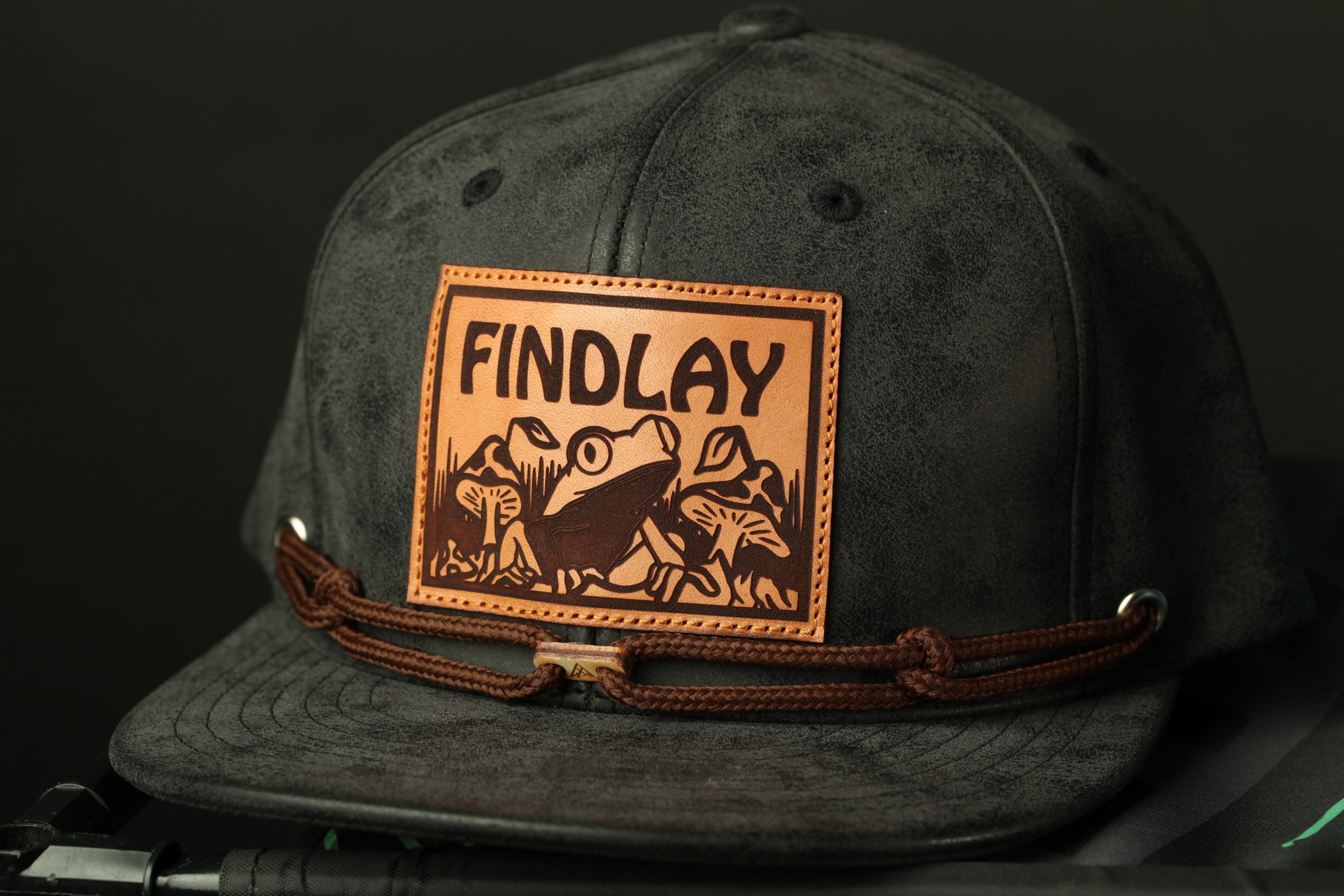 A black baseball cap with a brown leather patch on the front, featuring a frog and mushrooms.