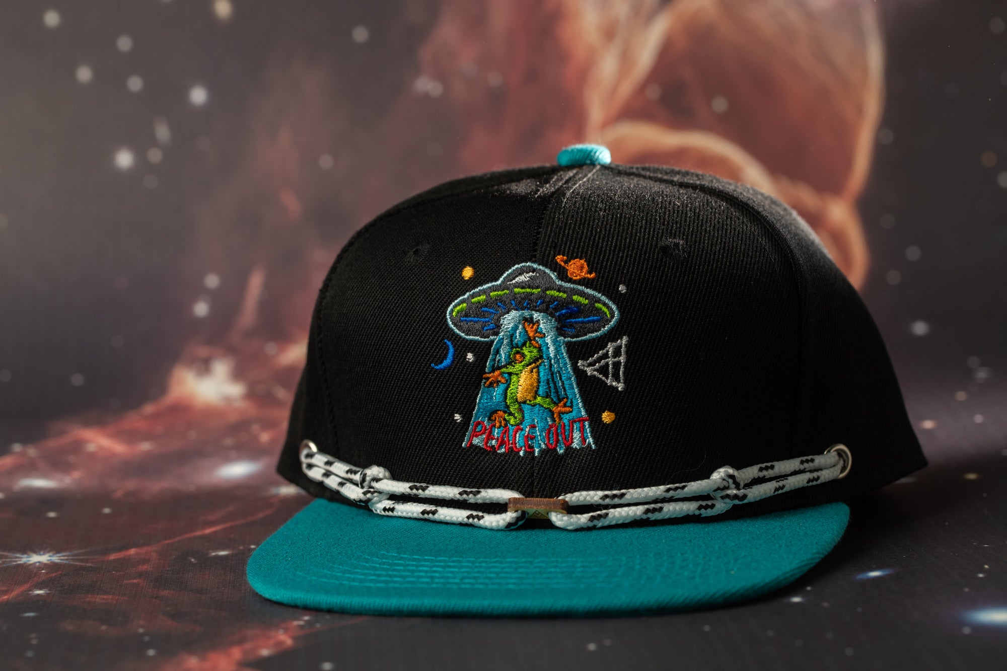 A black baseball cap with a colorful alien design and the text "PUNCH OUT" on it, placed on a dark background with a space-themed pattern.
