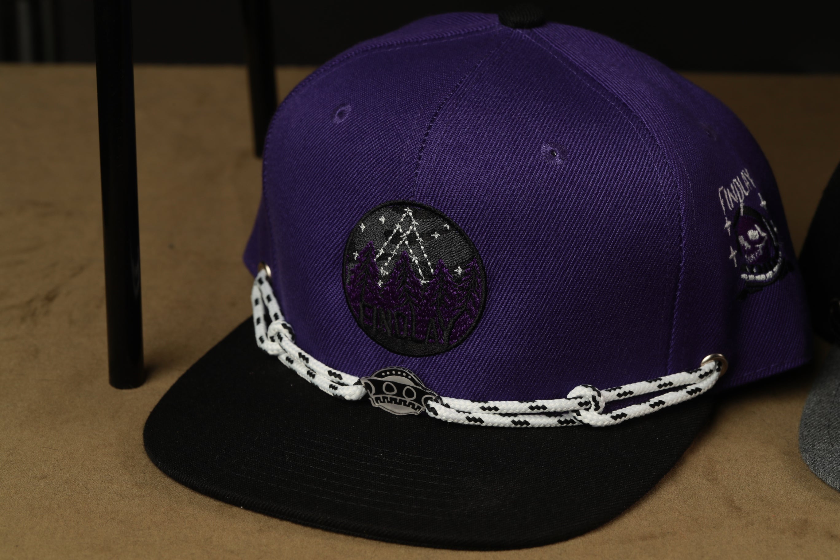 A purple baseball cap with a black brim and a white and black rope strap, placed on a wooden surface.