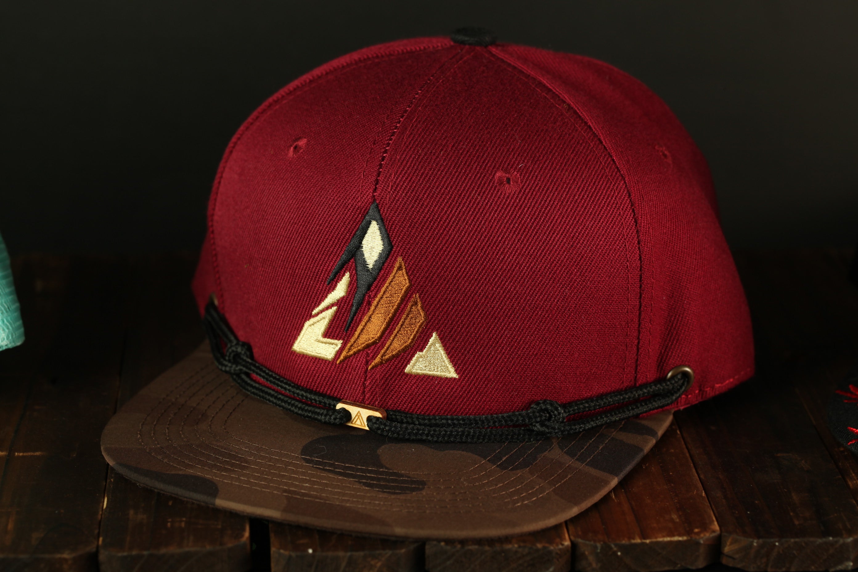 A maroon baseball cap with a camouflage pattern and a gold-colored logo on the front, placed on a wooden surface.