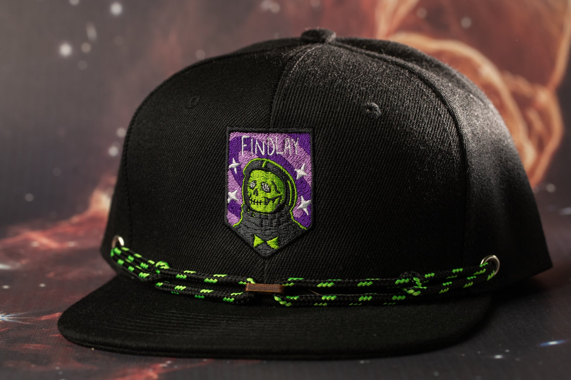 A black baseball cap with a purple and green patch featuring a skull and crossbones design, along with a green and black string.