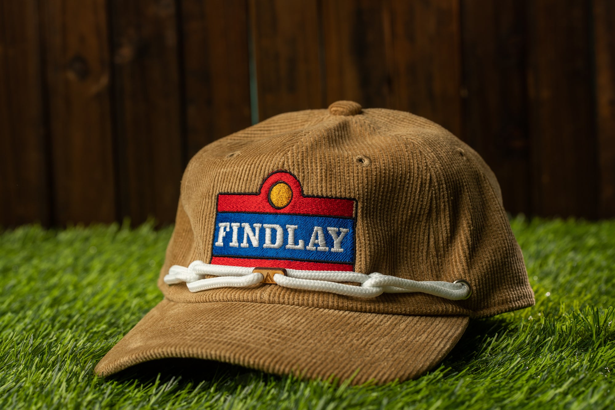 A brown baseball cap with a red and blue patch that says "FINDLAY" is placed on a grassy surface.