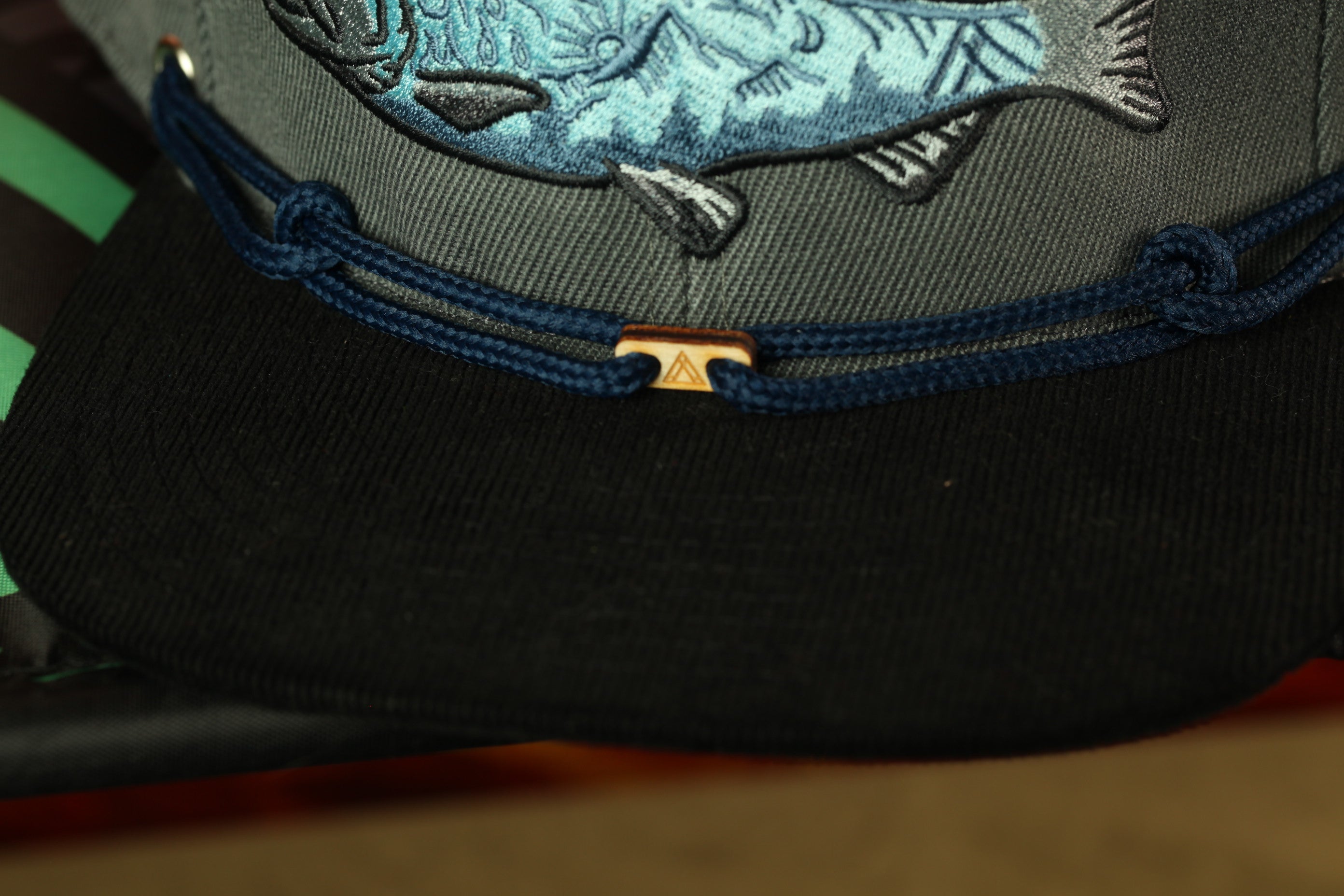 A close-up view of a black baseball cap with a fish embroidered on it, featuring a gold-colored metal clasp.