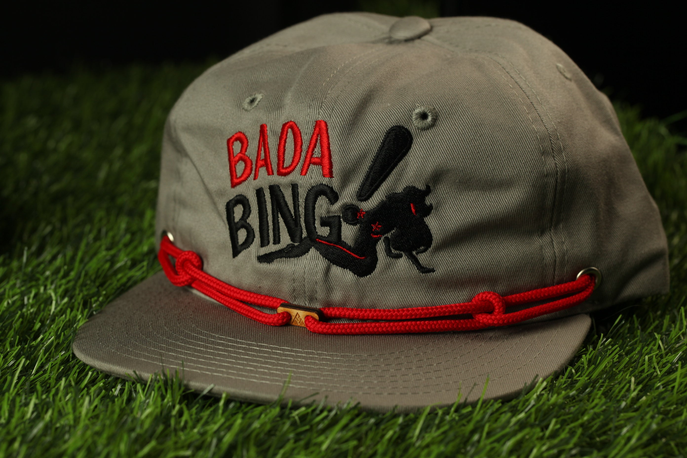 A gray baseball cap with the text "BADABING" printed on it, sitting on a grassy surface.