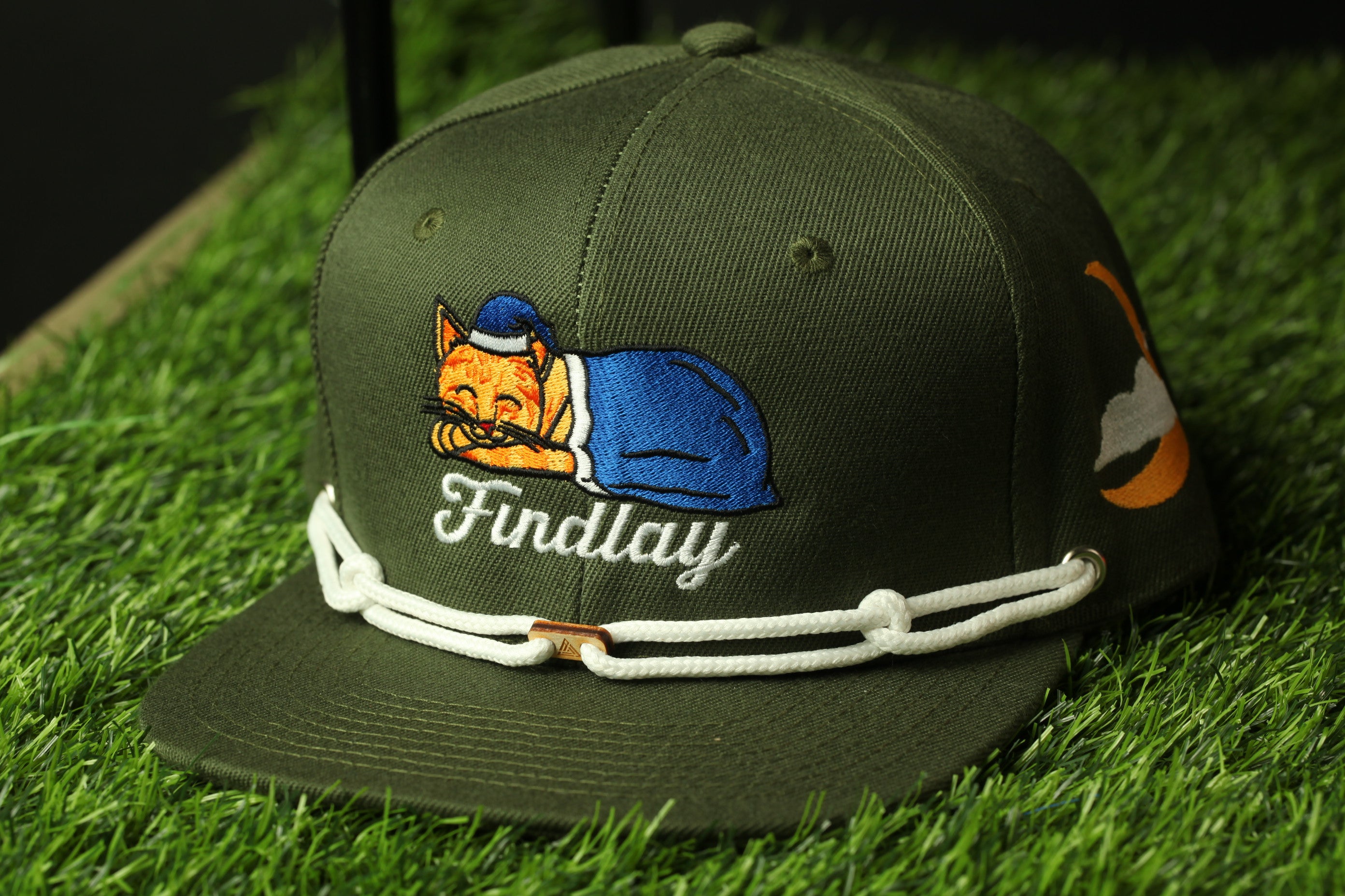 A green baseball cap with a cartoon cat design and the name "Findlay" printed on it, sitting on a grassy surface.
