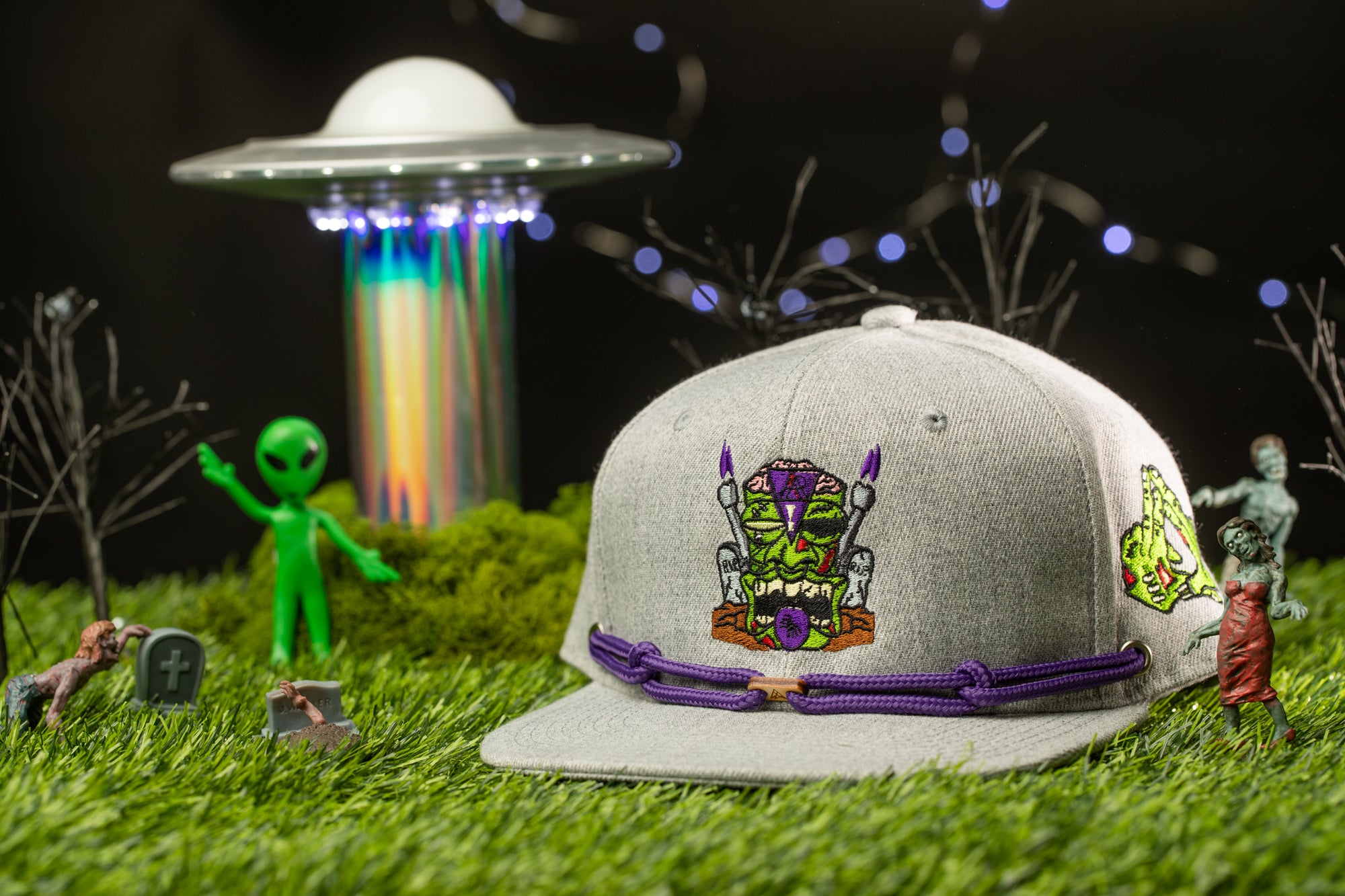 a gray baseball cap with purple accents, surrounded by various small toy figures and a green alien figure, all set against a dark background with a green alien structure in the background.