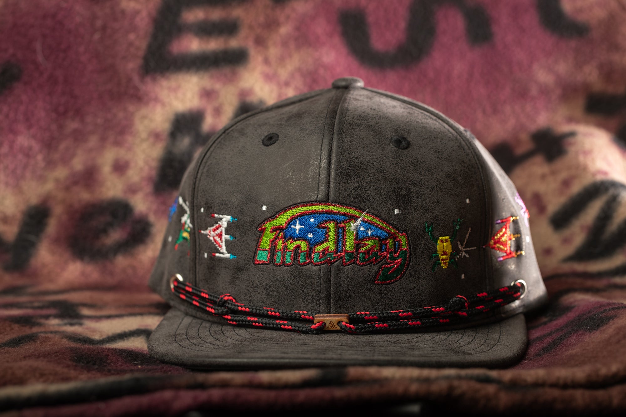 A black baseball cap with colorful embroidered designs and text on it, placed on a patterned fabric background.