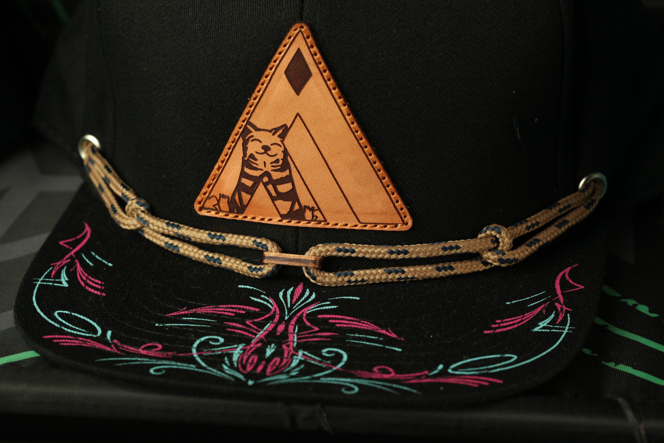 a close-up view of a black baseball cap with a unique design on the front. The cap has a triangular patch with a cat illustration, and a decorative pattern on the brim.