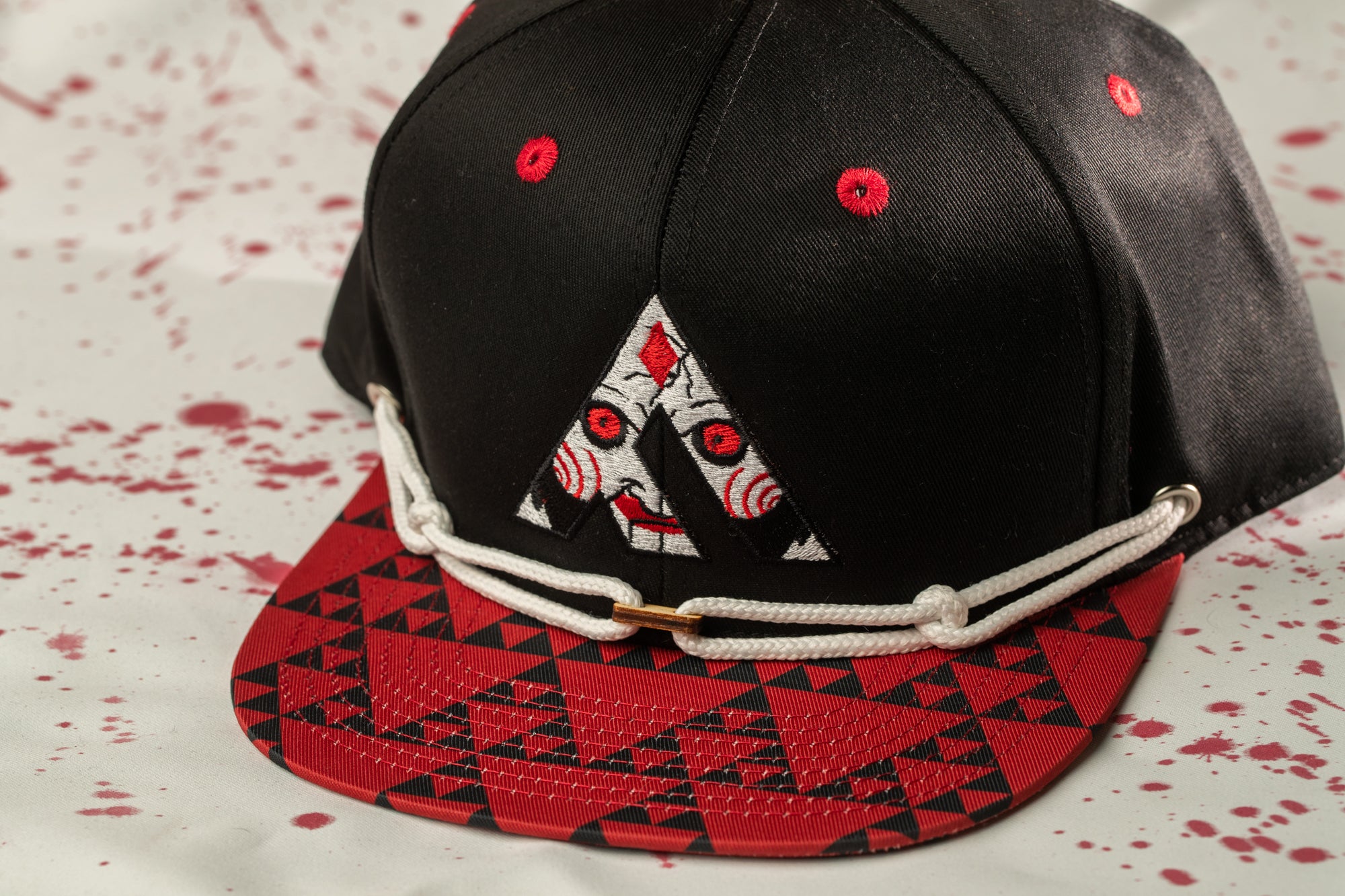 A black baseball cap with a red and black geometric pattern on the brim and a white string across the front.