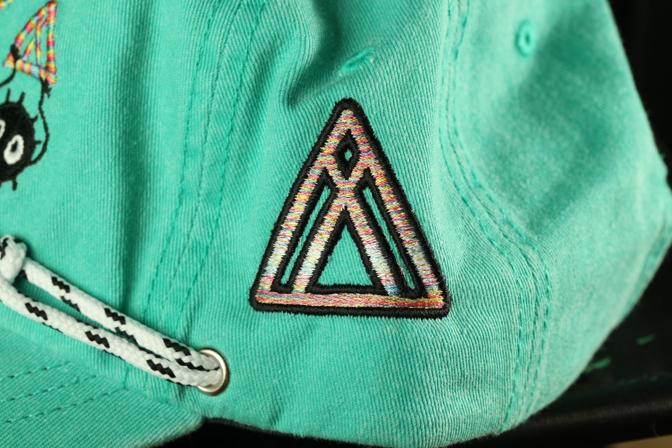 A close-up view of a turquoise baseball cap with a colorful triangle-shaped embroidered logo on the front.