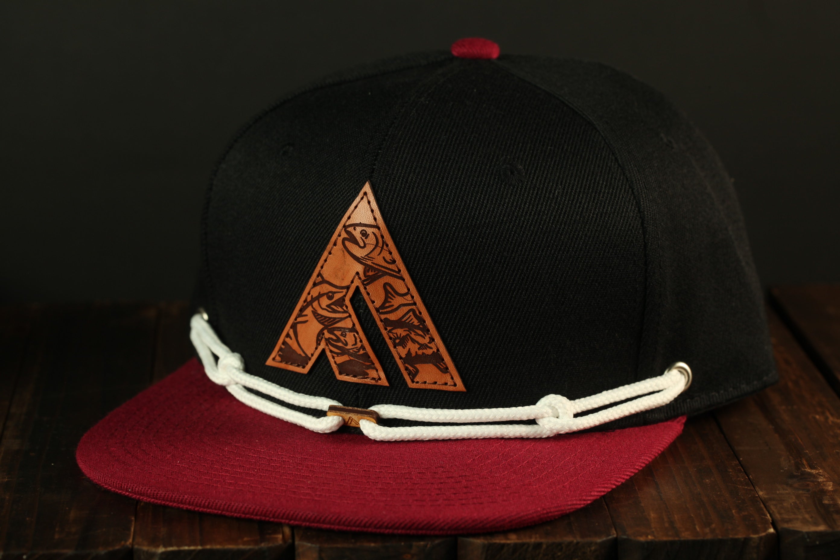 A black baseball cap with a red brim and a white lace trim, featuring a unique logo on the front.