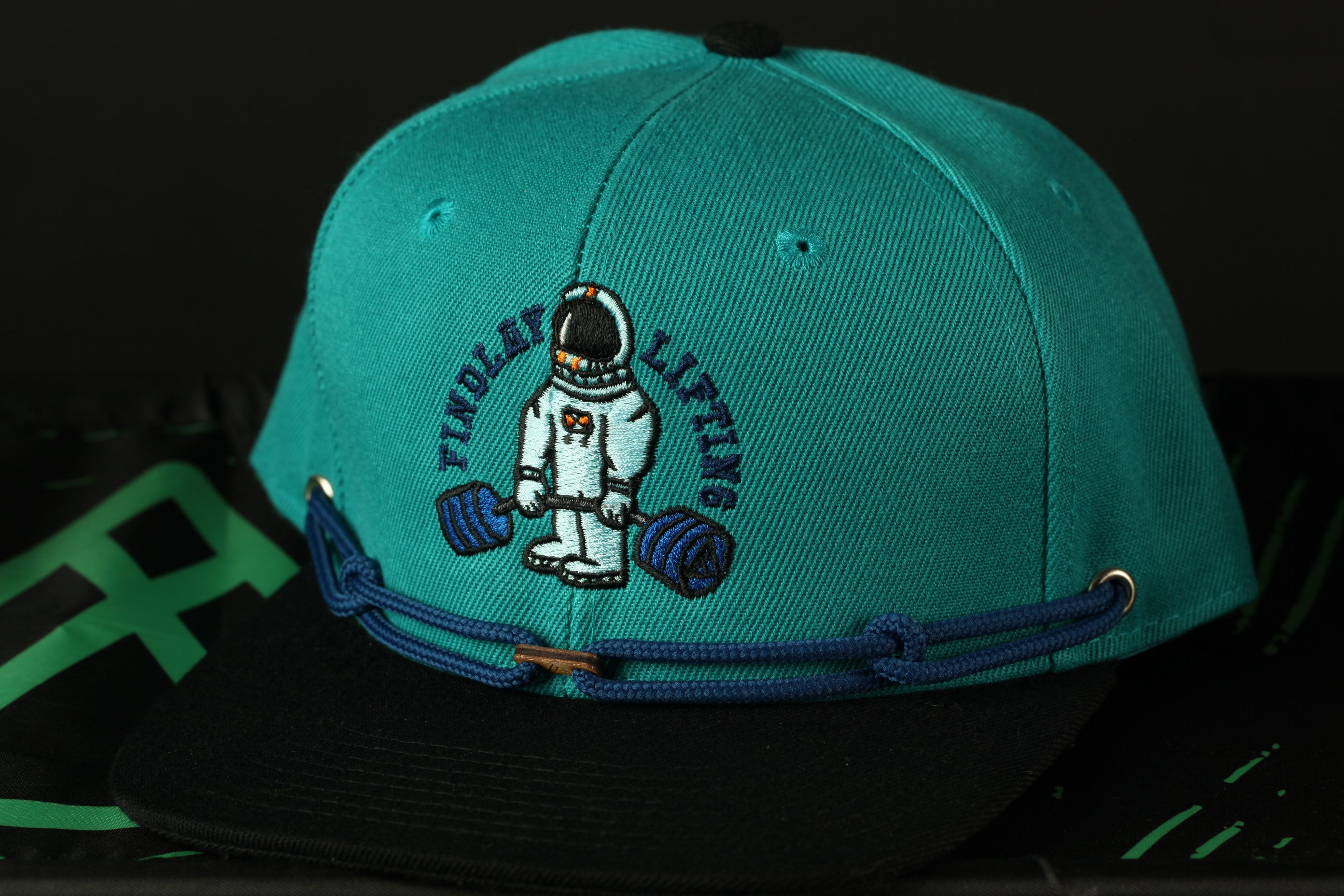 A turquoise baseball cap with a black brim and a blue strap, featuring an embroidered image of a person in a white suit holding a barbell.
