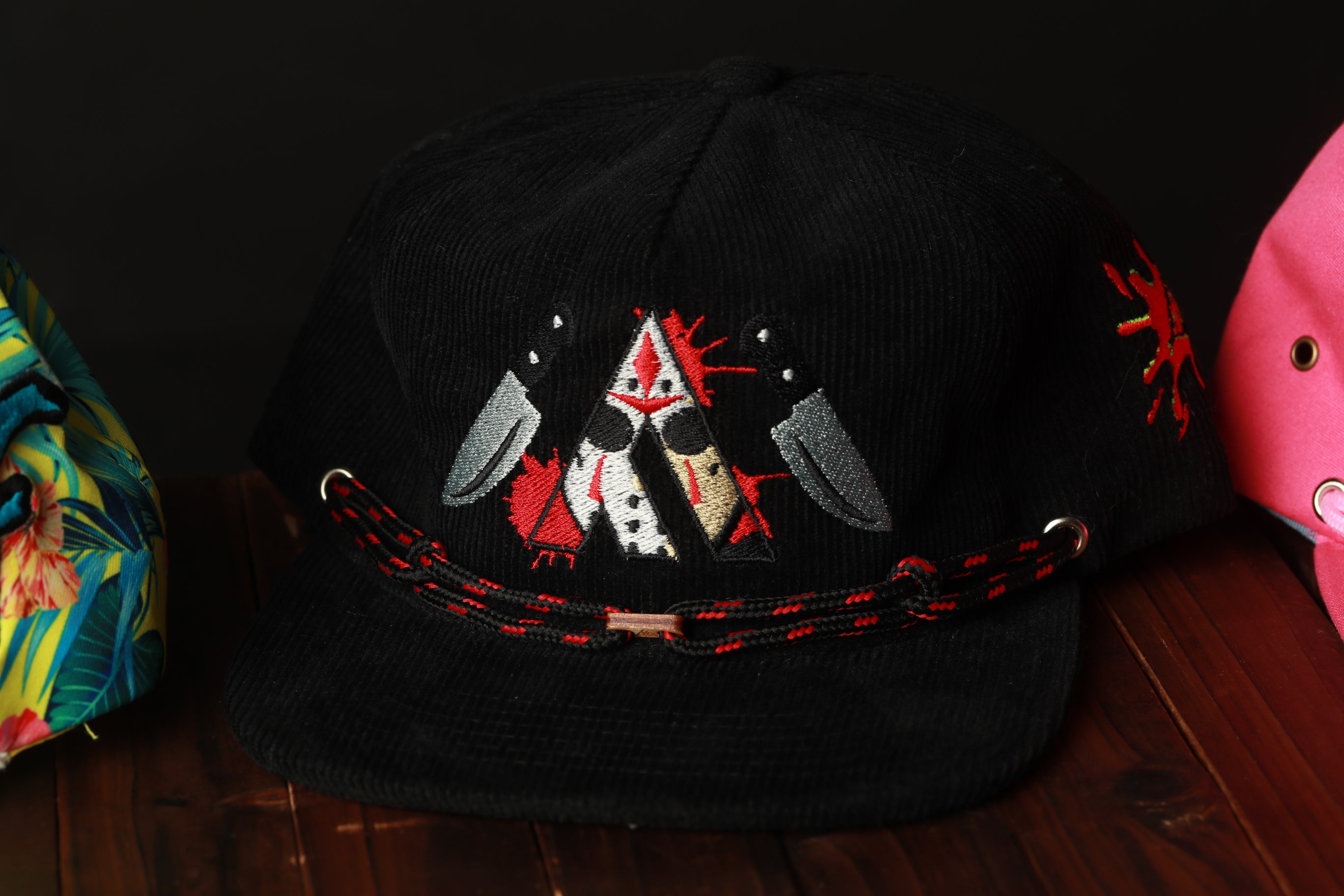 A black baseball cap with a red and white embroidered design on the front, featuring a stylized character with a knife.