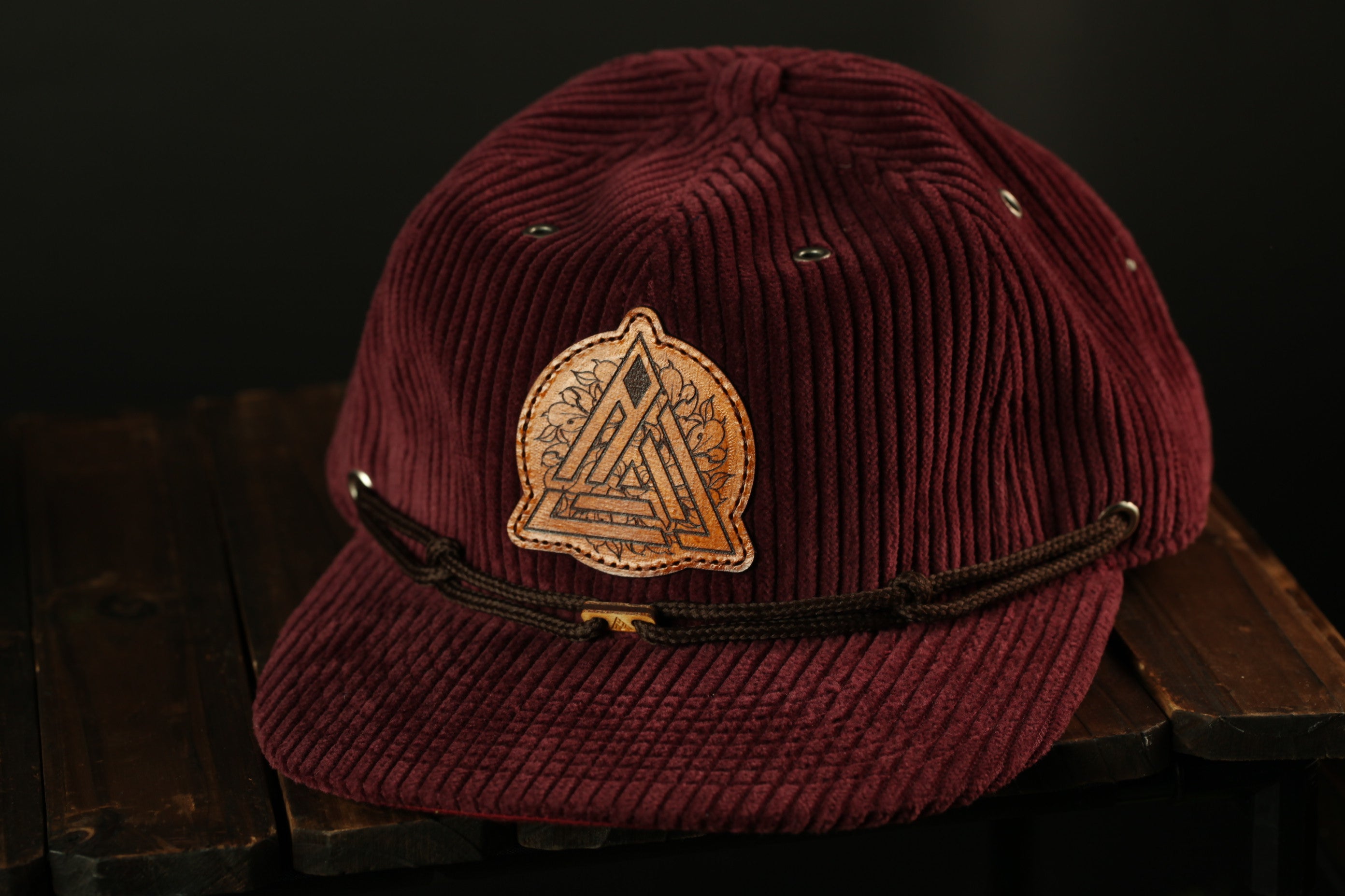 A burgundy baseball cap with a leather patch featuring a geometric design sits on a wooden surface.
