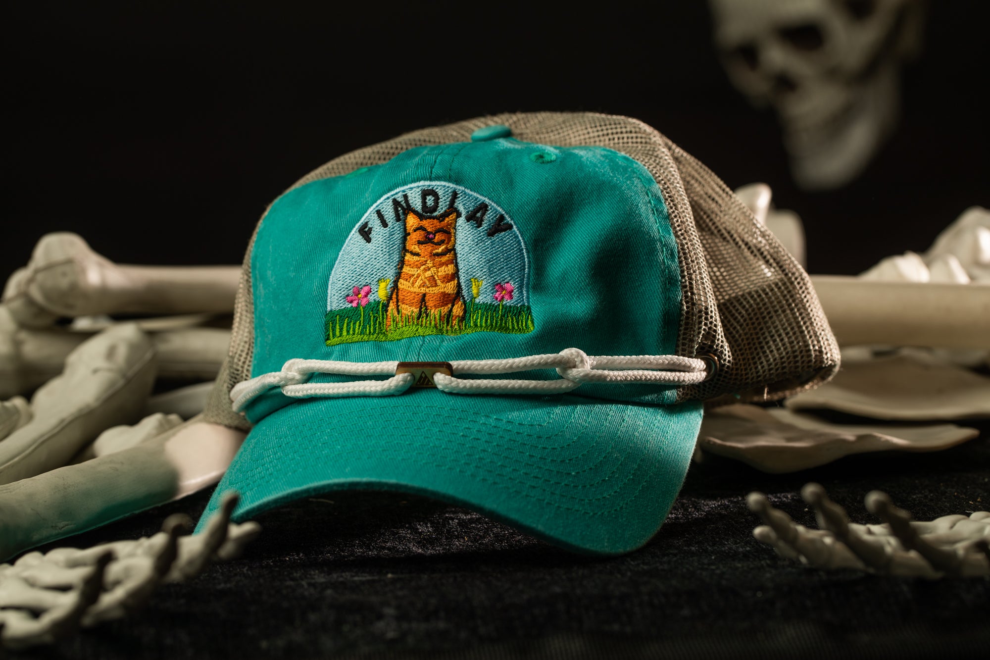 A turquoise baseball cap with a cat embroidered on it, placed on a surface with various objects, including bones and a skull.