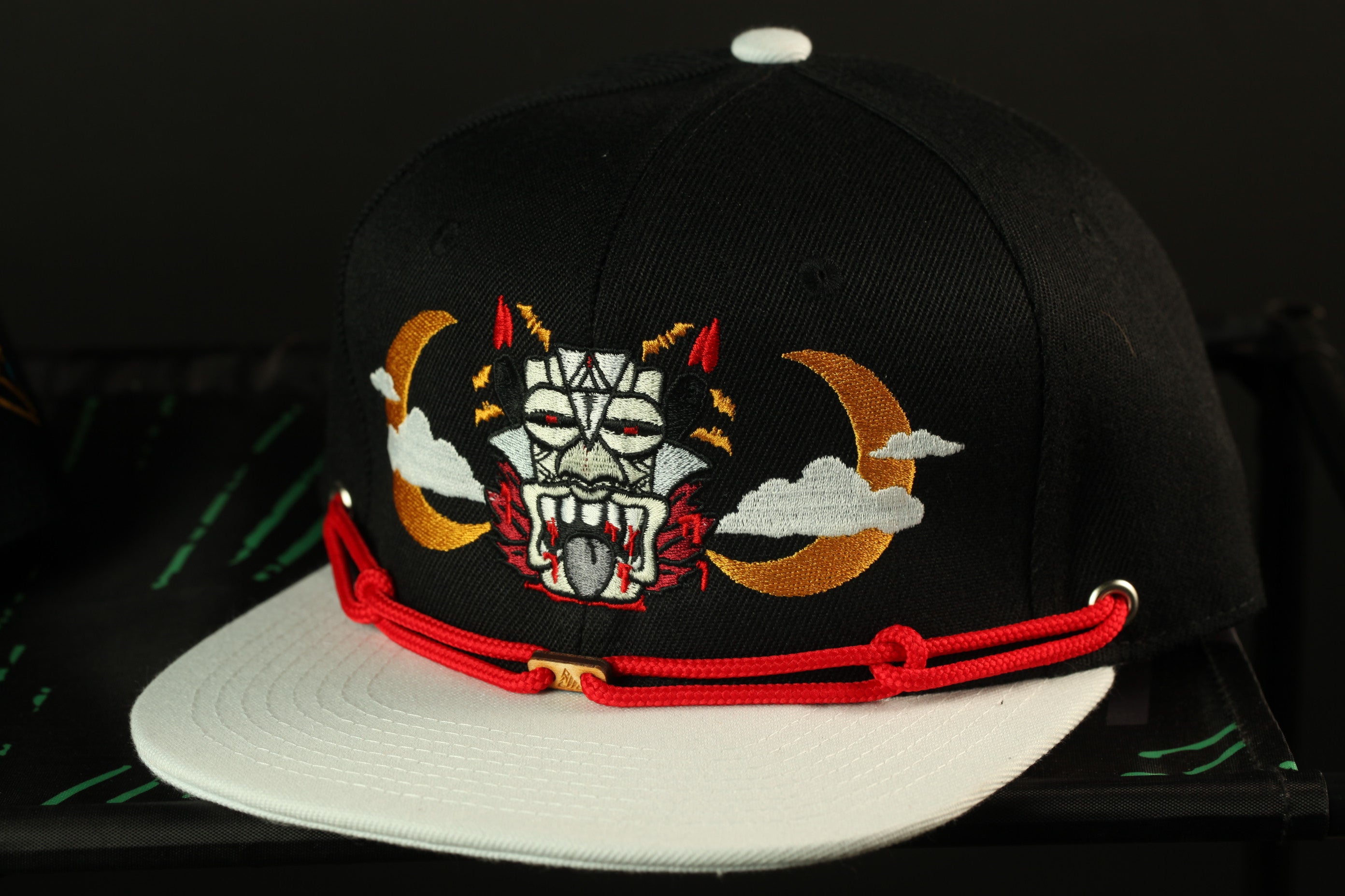 A black baseball cap with a white brim and a red string around the brim. The cap has an embroidered design on the front, depicting a mask with horns and a crescent moon.