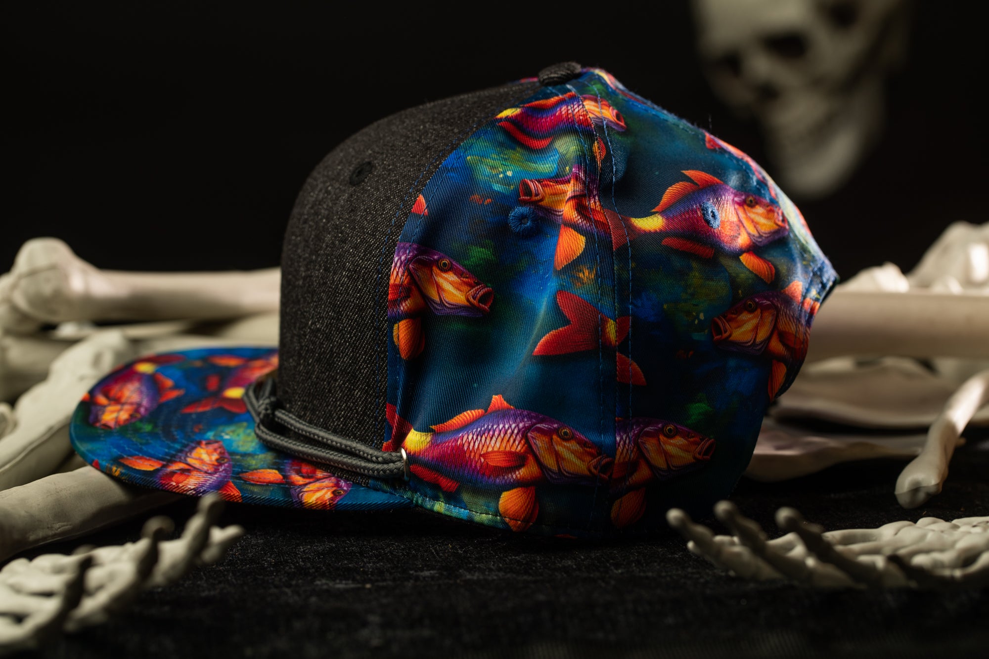 A colorful, patterned baseball cap with fish designs is placed on a dark surface, surrounded by skeletal bones.
