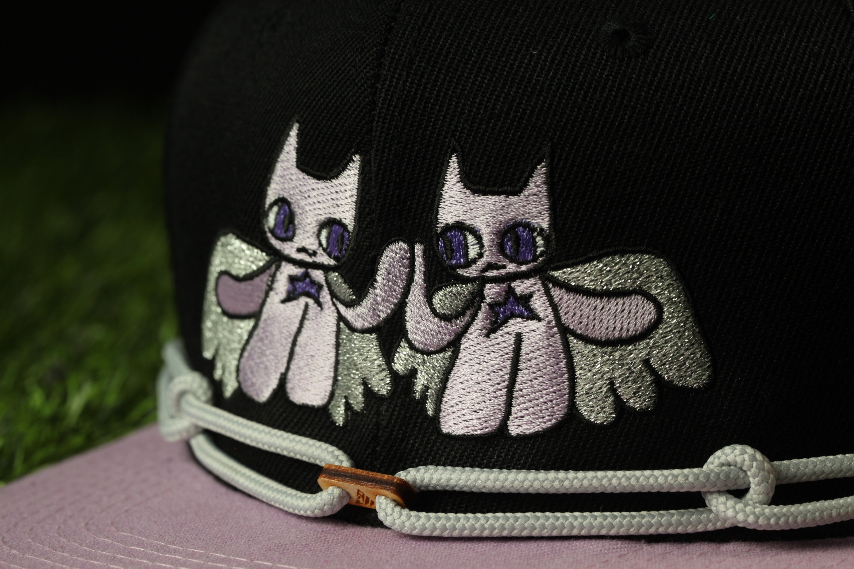 A close-up view of a black baseball cap with a unique embroidered design featuring two cartoon-like cats with wings.