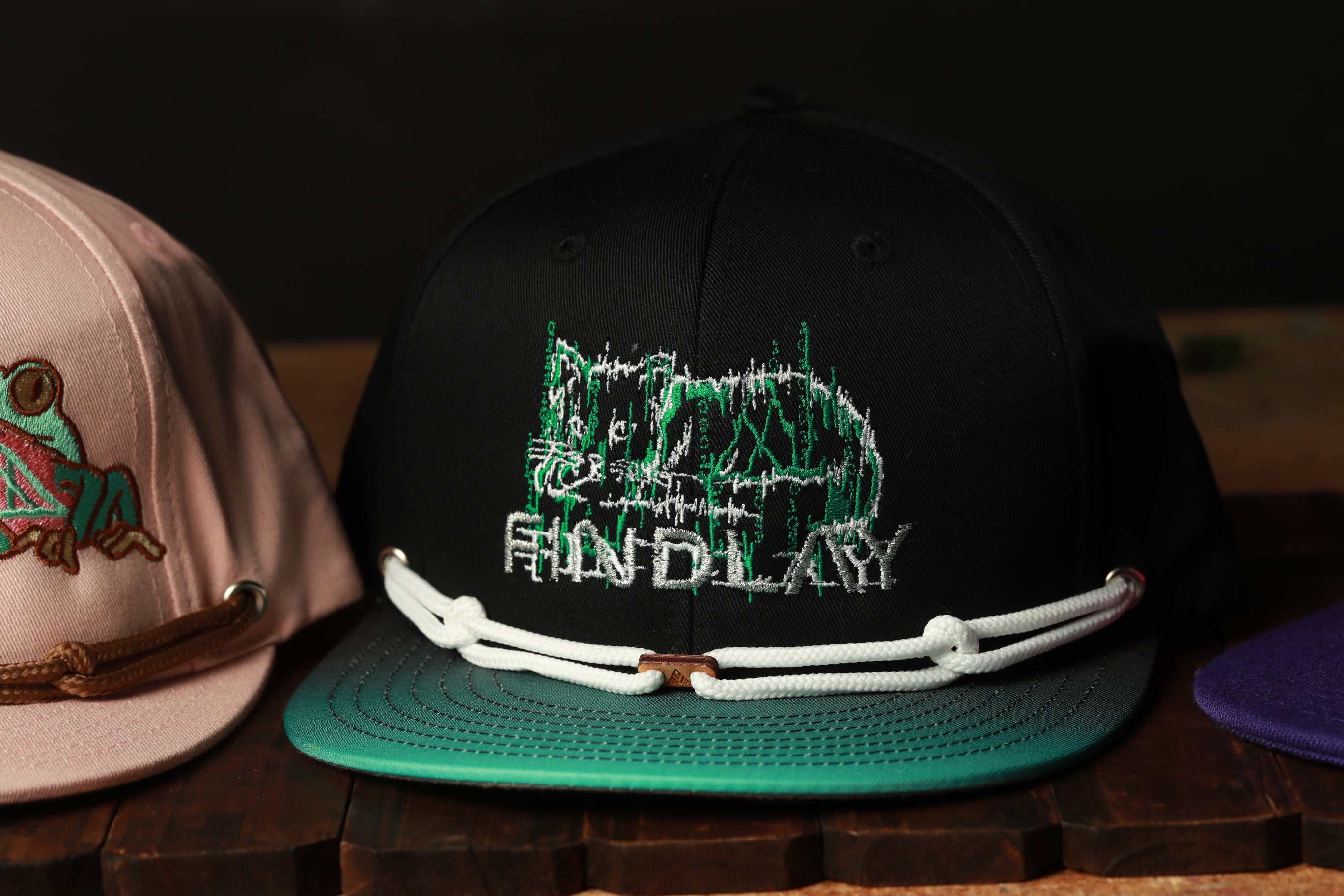 A close-up of a black baseball cap with a green cat design and a white rope strap, placed on a wooden surface.