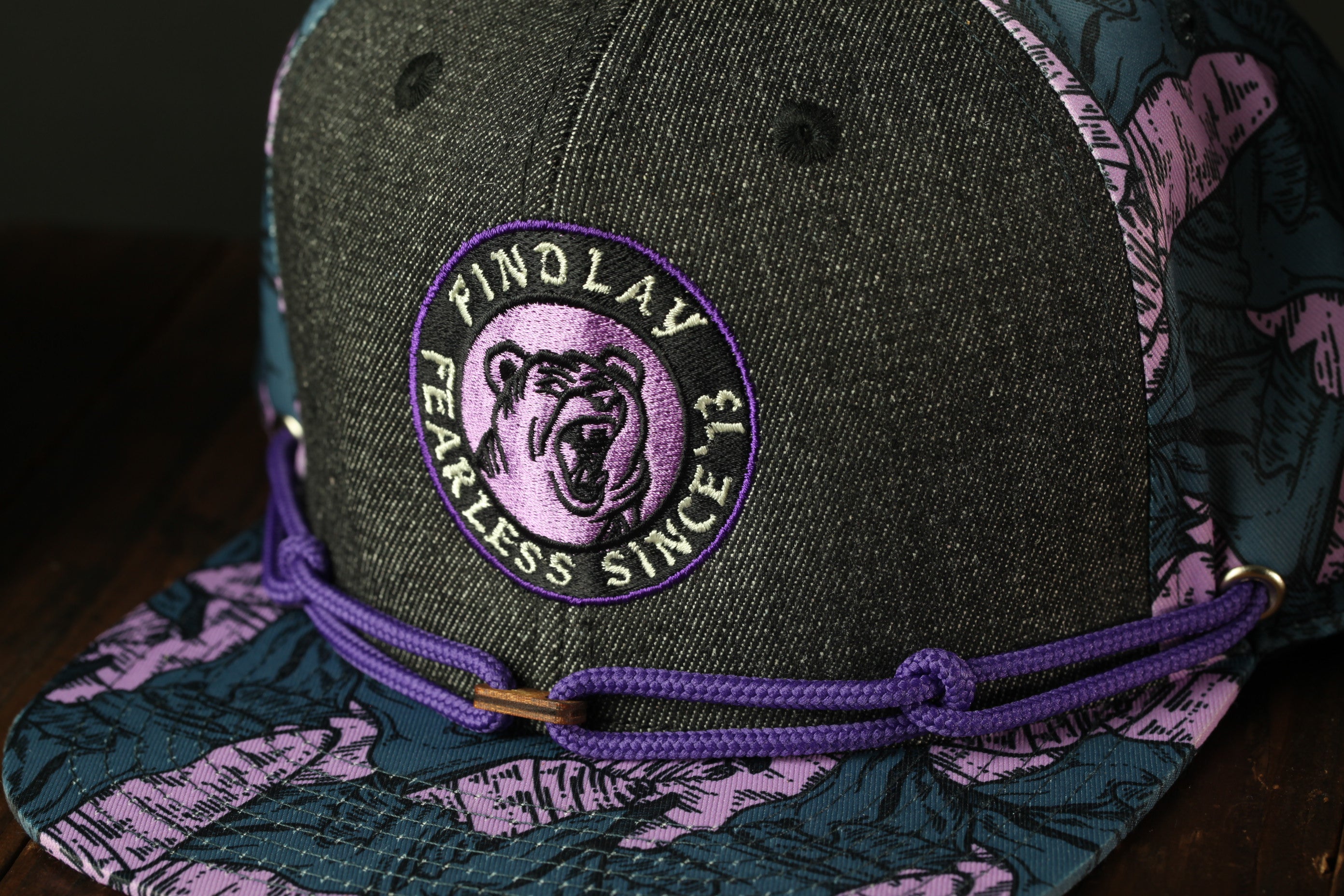 A close-up image of a black baseball cap with a purple and pink floral pattern, featuring a circular logo with a bear's face and the text "Finnlay Fearless Since 13".