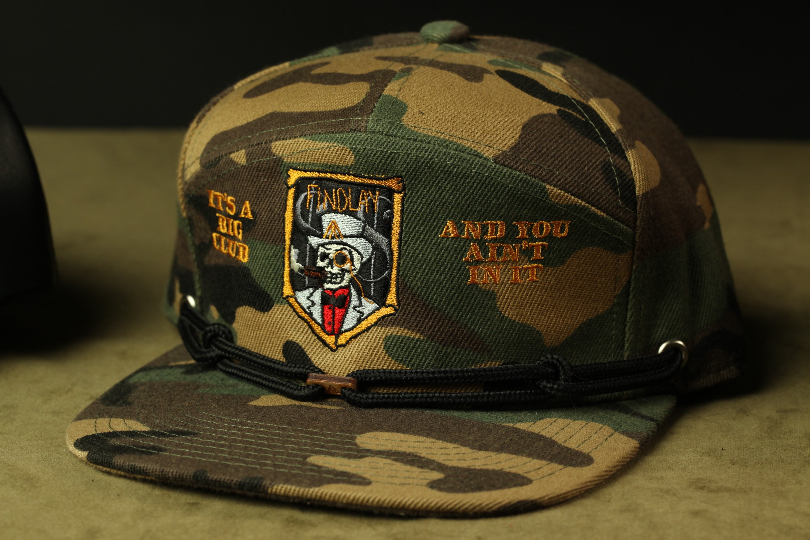 A camouflage baseball cap with a skull and crossbones design, and text on the front.