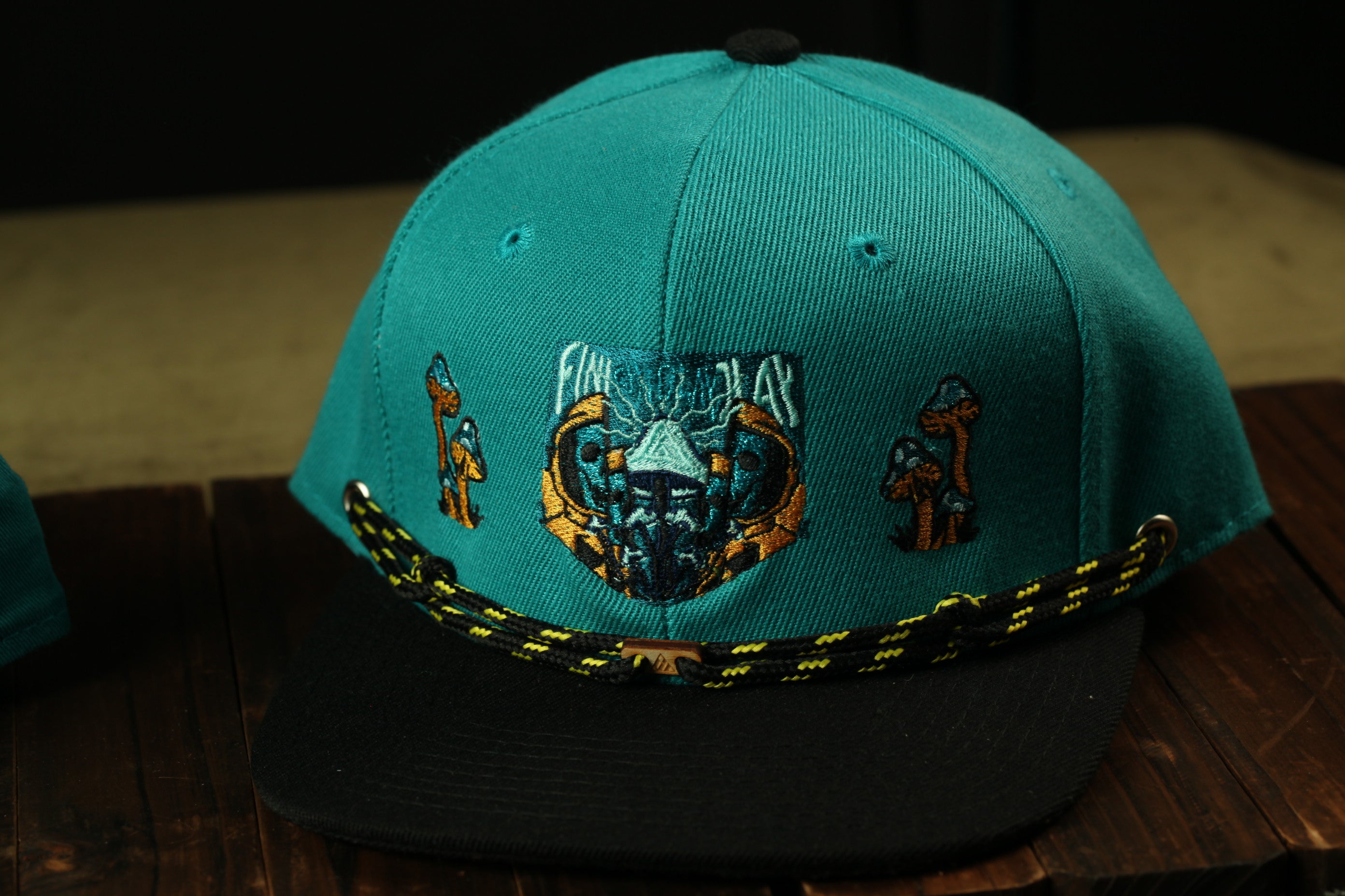 A turquoise baseball cap with a black brim and yellow and black braided strap, featuring a graphic design on the front.